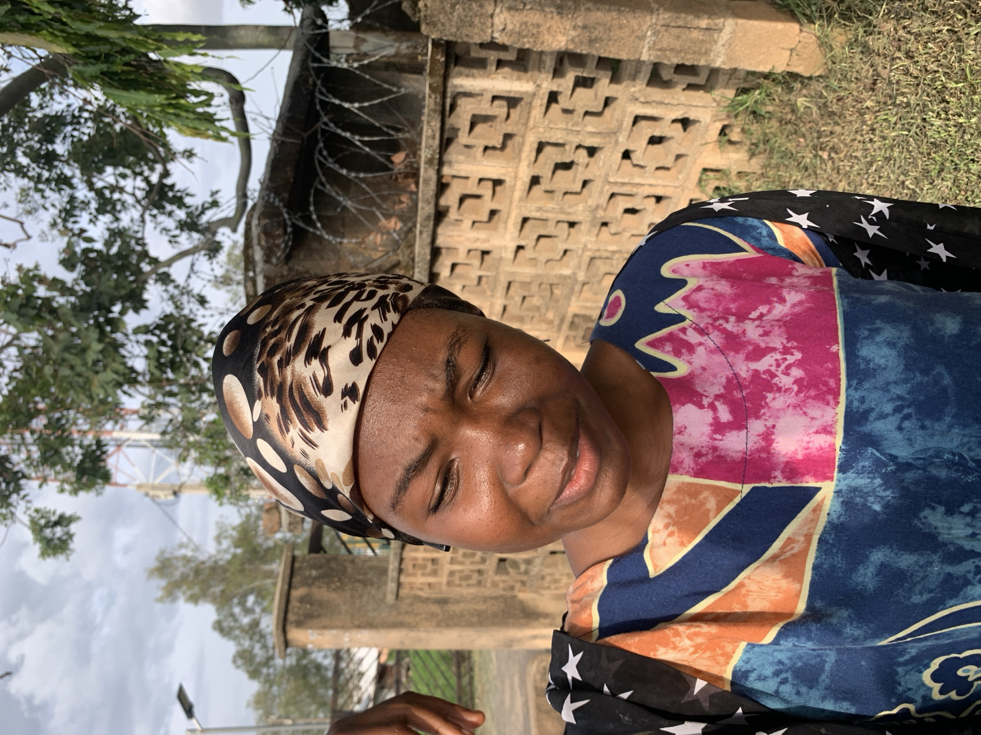 A medium shot captures a young Black woman outdoors, her head adorned with a leopard-print headscarf. Her eyes are cast downwards, and her lips are slightly parted. She wears a colorful, patterned garment with broad sections of blue, orange, pink, and yellow. A black sleeve with white stars is visible on her left side. In the background, a concrete wall with a decorative, cutout pattern stretches across the frame, topped with barbed wire. Beyond the wall, green foliage from trees is visible, along with a distant cellular tower. The scene appears to be set in a moderately urban environment, possibly within the city of Jos, Nigeria, given the architectural and atmospheric cues. The lighting suggests it is daytime, with the sun casting subtle shadows.