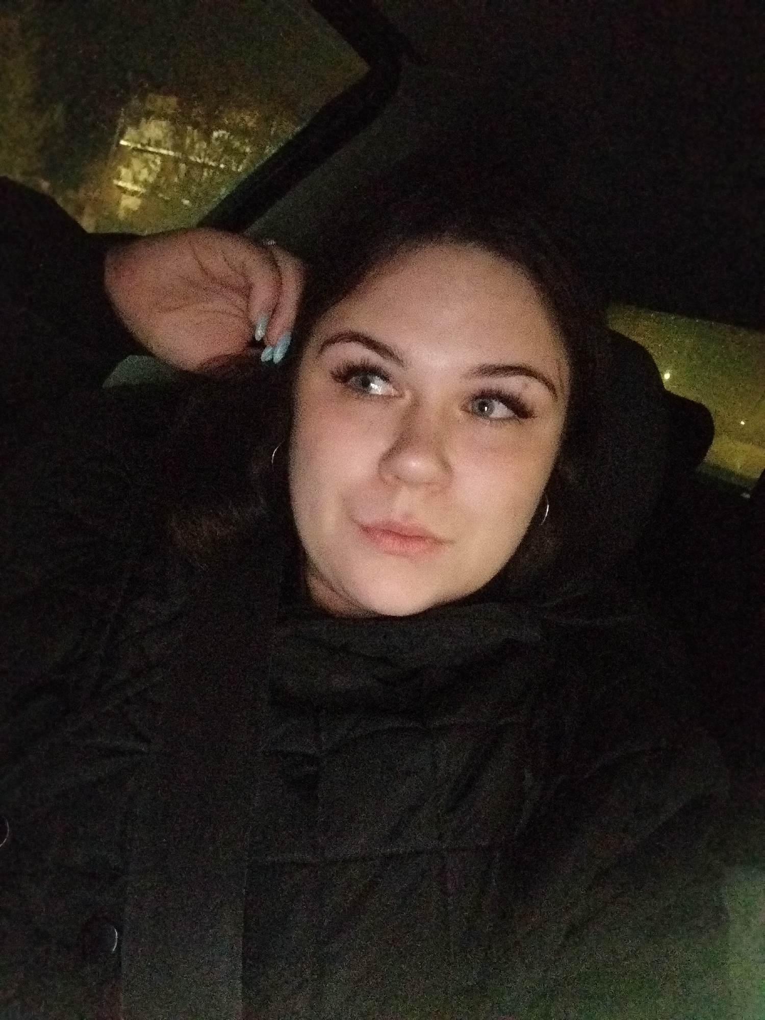 A woman is in a vehicle at night, likely a car. She has dark hair, light skin, and light-colored eyes. She is wearing a dark jacket with a high collar. Her left hand is raised to her head, with light blue nail polish visible. The background shows blurred lights, potentially from the city. Given the prompt, it's possible the location is in Chaikovsky, Russia. The interior of the vehicle appears dark, with the woman positioned against what looks like the headrest of a seat.
