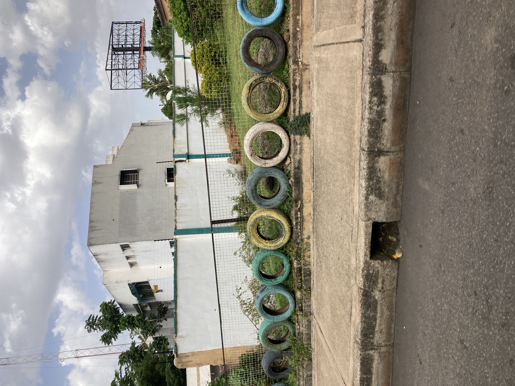 The image depicts a street scene with a white building and a row of colorful tires bordering a sidewalk. The building appears to be multi-story and has several windows. A large, metal billboard structure is visible above and to the left of the building. The tires, painted in various shades of blue, yellow, green, and white, are arranged in a line and appear to be used as a decorative border or barrier along a chain-link fence. Vegetation, including trees and bushes, is present behind the fence and tires. The sidewalk is made of concrete, and next to it is an asphalt road. A drain is visible in the curb. The sky is cloudy, suggesting daytime with overcast weather. The scene appears to be in an urban or semi-urban environment. There is no visible text in the image. The overall impression is one of an outdoor, public space, possibly in Jos, Nigeria, given the provided location context.