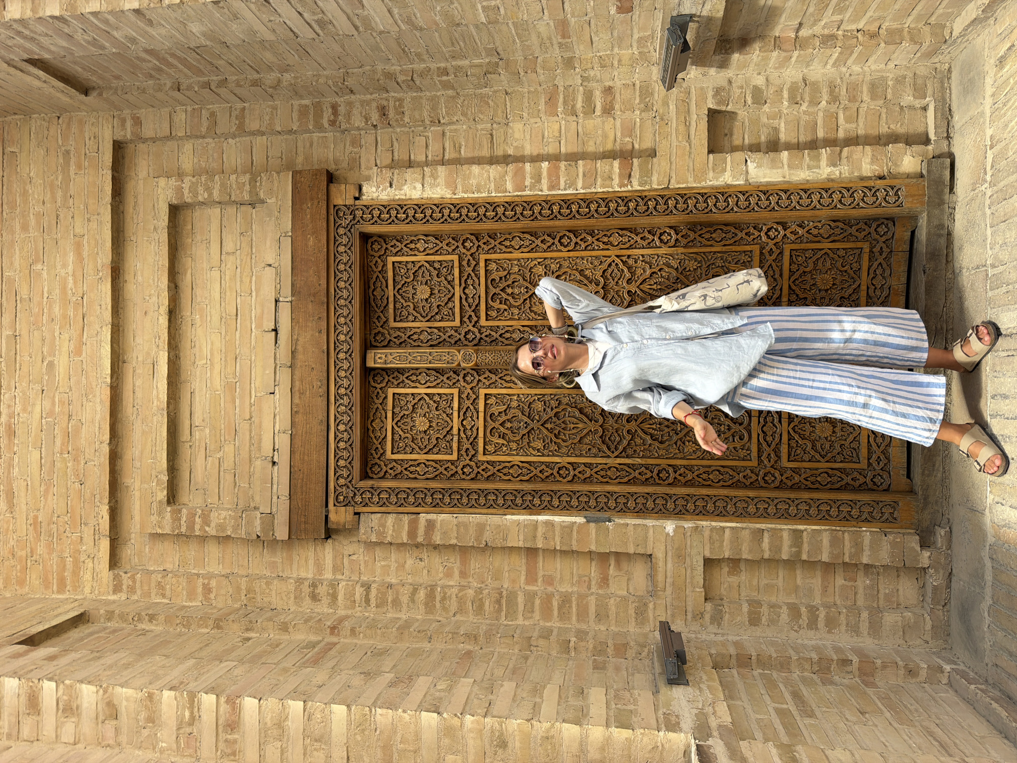A young woman stands in front of a large, intricately carved wooden door. The door is set within a brick wall. The woman is wearing a light blue collared shirt, blue and white striped wide-leg pants, and tan sandals. She has on sunglasses and her hair is pulled back. She is holding a tote bag over her left shoulder and her right hand is resting on the back of her head. The brick wall appears to be made of light-colored bricks and has some decorative brickwork around the door. The scene suggests an outdoor location, possibly an architectural site or a historical building in Samarkand, Uzbekistan, given the style of the door and brickwork.
