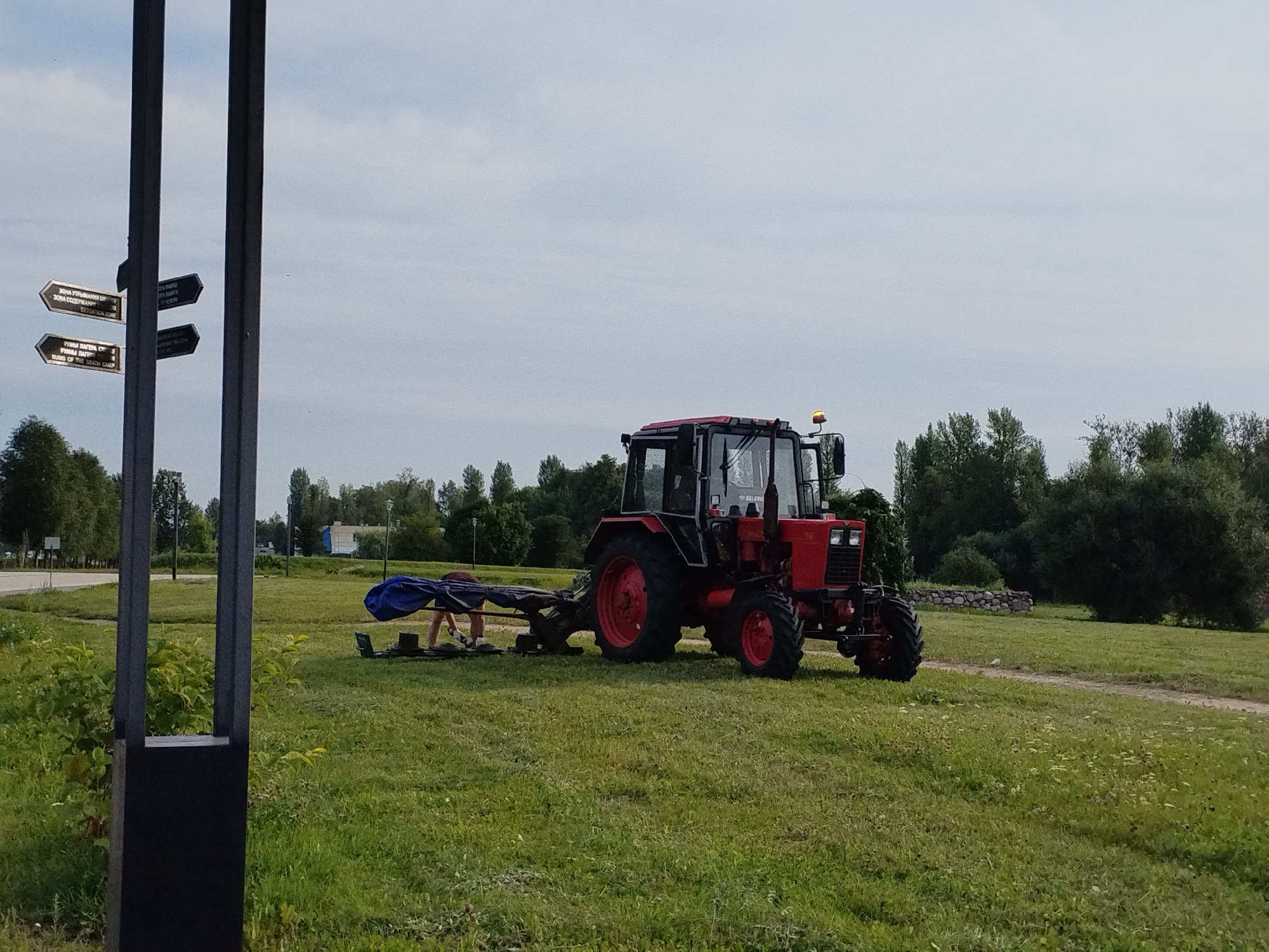 In the city of Minsk, Belarus, on a grassy field under a cloudy sky, a red tractor with a black cabin and yellow flashing light is parked. Attached to the tractor is an implement with a blue tarp draped over it. A person, wearing a dark shirt and light shorts, is positioned behind the tractor, appearing to be working with the implement. To the left of the tractor, a dark metal post with several directional signs is visible. The signs, written in Cyrillic and English, indicate locations such as "DETENTION ZONE" and "RUINS OF THE DEATH CAMP." The environment suggests a park or open area with trees in the background.