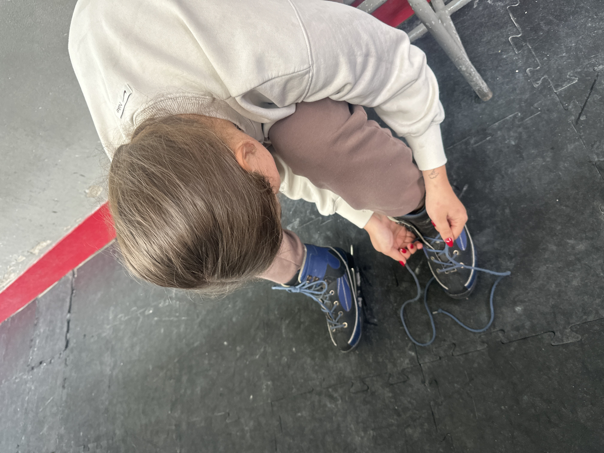 A young person with brown hair pulled back is kneeling on a black interlocking mat floor, tying the laces of a blue and black athletic shoe. The shoe is one of a pair, with the other shoe on the person's left foot. The person is wearing a light-colored crewneck sweatshirt and brown sweatpants. A red stripe runs diagonally across the upper left corner of the image. The scene is indoors, and the floor appears to be a gym or training facility. The person's hands are visible, with red nail polish on their fingernails, as they manipulate the shoelaces. A small tattoo is visible on the person's left forearm.