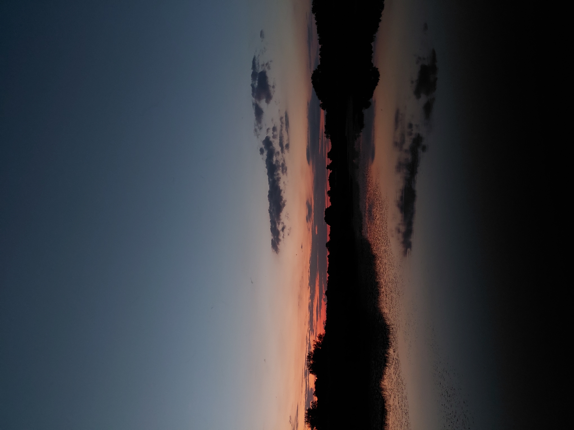 The image depicts a serene natural landscape at dusk in the city of Ustyvytsia, Ukraine. The sky transitions from a deep twilight blue at the top to hues of orange and pink near the horizon, indicating the setting sun. A silhouetted tree line forms a dark band across the middle ground, with scattered clouds visible in the sky. Below the tree line, a body of water reflects the colors of the sky and the silhouette of the trees. The foreground is dominated by the dark surface of the water, with subtle ripples and reflections. The overall scene conveys a sense of tranquility and the calm beauty of nature at the end of the day. There are no people, animals, or man-made structures visible in the image.