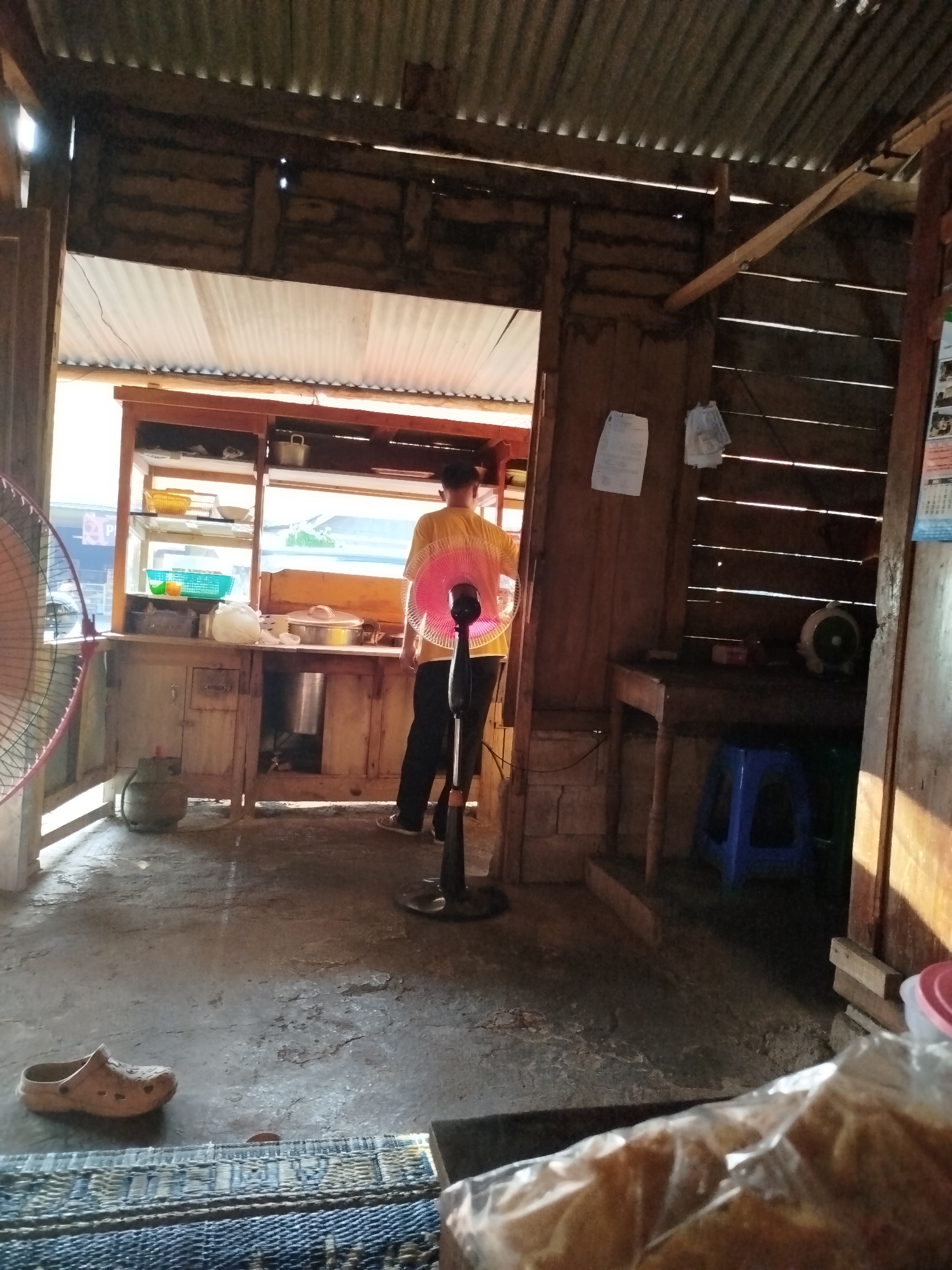 The image depicts the interior of a humble food stall in the city of Pojok, Indonesia. The stall is constructed with wooden planks and a corrugated metal roof, suggesting a simple, rustic setting. Sunlight streams in from an opening, illuminating the dusty concrete floor and casting long shadows.
A lone individual, wearing a yellow shirt and black pants, stands facing away from the camera, attending to the food preparation area. In front of them is a wooden counter displaying various items, including what appear to be kitchen utensils and containers. A pink-bladed electric fan is positioned prominently in the foreground, suggesting a hot climate.
To the left, another large electric fan is visible, partially obscuring a gas cylinder. The floor is bare concrete, marked with cracks and wear. A single brown sandal rests on the floor near a woven mat. In the background, a blue plastic stool is tucked under a wooden table. The overall scene conveys a sense of everyday life and commerce within this Indonesian city.