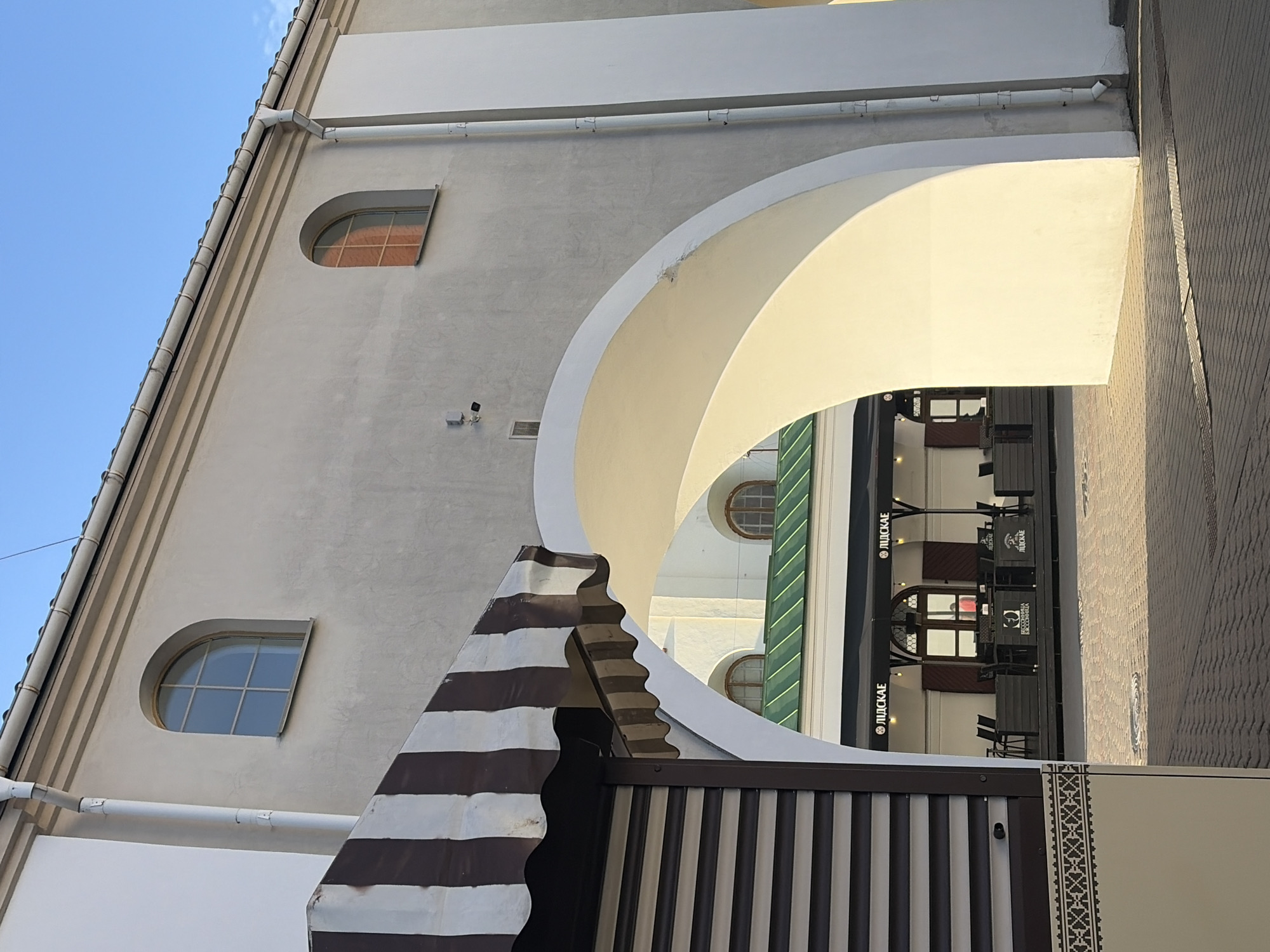 The image depicts an outdoor scene in Minsk, Belarus, likely during daylight hours given the clear blue sky and sunlight casting shadows. In the foreground, a portion of a building with a striped awning and closed shutters is visible on the left. The awning features alternating brown and white stripes.
The central element of the image is a large, smooth, white archway that leads into a courtyard or open space. Through the arch, a cafe or restaurant with outdoor seating can be seen. The cafe has black awnings with the Cyrillic text "ЛІДСКАЕ" written on them. Several tables and chairs are set up on a dark wooden deck. The architecture of the building behind the cafe features multiple arched windows with dark frames.
The building surrounding the archway is painted a light, neutral color and has a downspout running vertically along its side. The ground in the foreground and leading into the archway is paved with brick or cobblestone. The overall impression is one of an urban setting, possibly a historical or pedestrianized area, with a focus on architecture and a dining establishment.