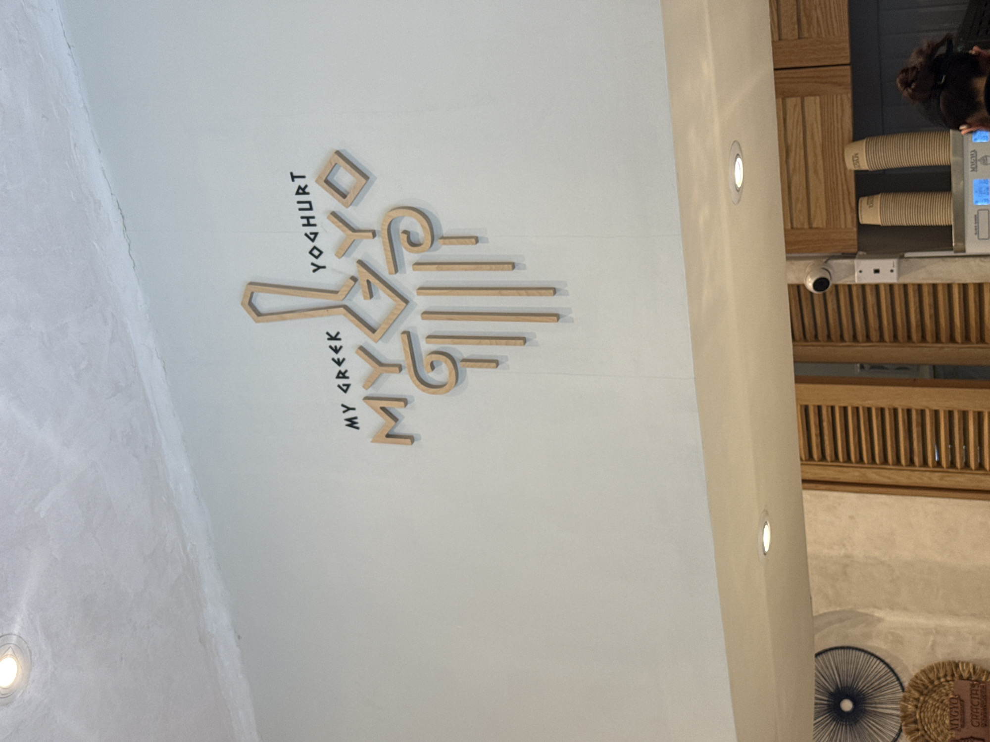 The image shows a wall with a logo that reads "MY GREEK YOGHURT" in a stylized design. The letters are made of wood or a similar material. The background is a white wall. To the right of the wall, there is a section with wooden slats, and a surveillance camera is visible. There are also objects that appear to be paper cups and what seems to be a payment terminal. People are partially visible in the upper right corner. The photo was taken indoors. Based on the logo, it's likely taken in Mexico City, Mexico.