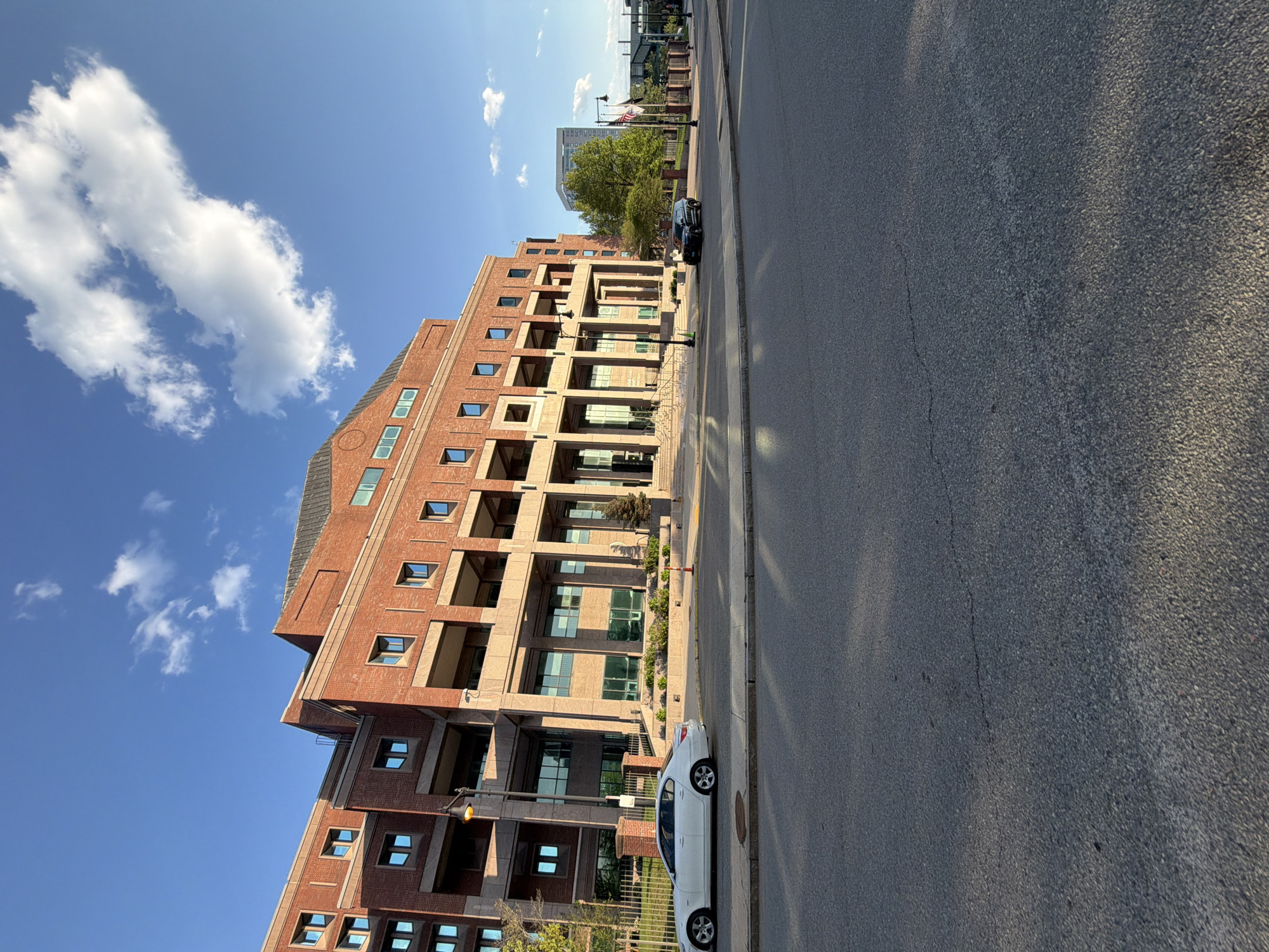 The photograph depicts a street scene in Boston, Massachusetts, USA, featuring a large brick building with multiple stories and numerous windows, under a blue sky with scattered clouds. The building has a distinctive architectural style with protruding sections and what appear to be balconies or recessed areas. A street runs alongside the building, with parked cars visible. Light poles are positioned along the sidewalk. In the distance, other buildings and trees are discernible, along with what seems to be an American flag. The road surface shows wear and tear with cracks visible in the pavement. The overall lighting suggests it is daytime, likely in the late morning or early afternoon, with shadows cast by the building.