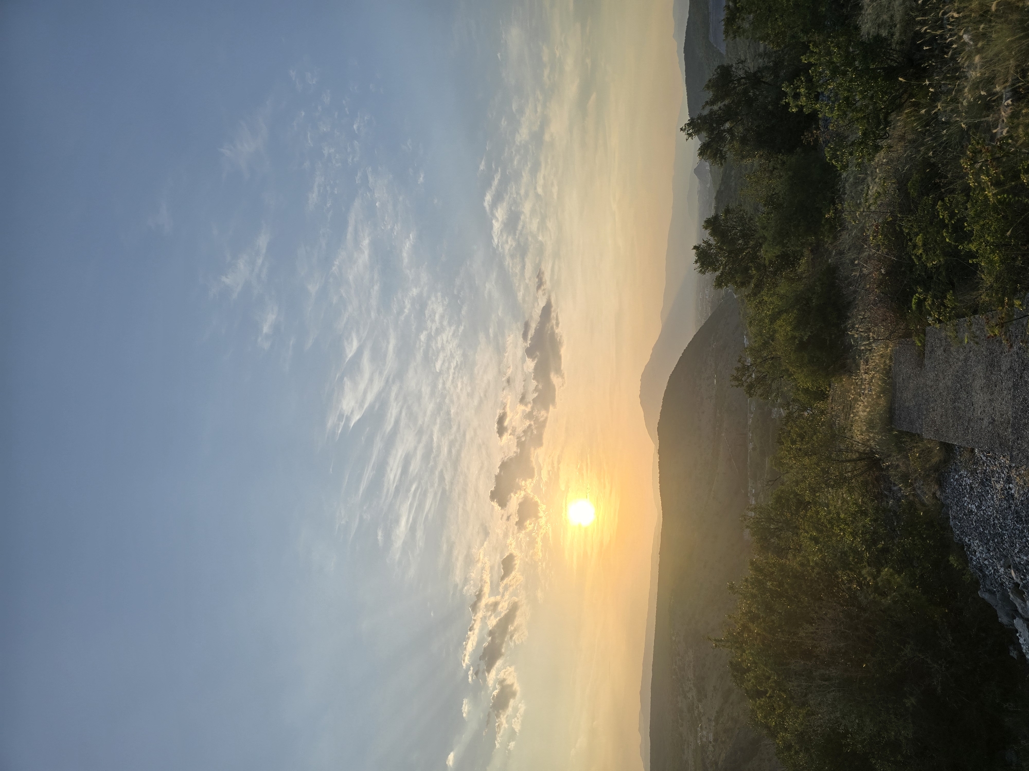 This is a vertical, eye-level, full-shot photograph taken at sunset from a high vantage point overlooking the city of Podgorica, Montenegro. The sky dominates the upper half of the image, transitioning from a pale blue at the top to a warm yellow and orange near the horizon, indicative of the setting sun. Wispy, white clouds are scattered across the sky, some catching the golden light of the sun.
The sun itself is a bright, luminous orb positioned slightly to the left of the center in the mid-upper portion of the frame. Rays of sunlight emanate outwards, creating subtle atmospheric effects.
Below the sky, a series of rolling hills and mountains recede into the distance, their contours softened by the haze of sunset. The closest hills are covered in dense, green vegetation. In the middle ground, hints of the city of Podgorica are visible, with buildings and structures partially obscured by the landscape and the lighting conditions.
The foreground of the image features a rough, rocky terrain interspersed with dry grasses and shrubs. A concrete pathway or step is visible in the lower right quadrant, leading downwards. Several trees with lush green foliage are scattered throughout the foreground and middle ground, their leaves illuminated by the golden light of the setting sun. The overall scene evokes a sense of natural beauty and tranquility at the end of the day, with the city nestled within the expansive landscape.