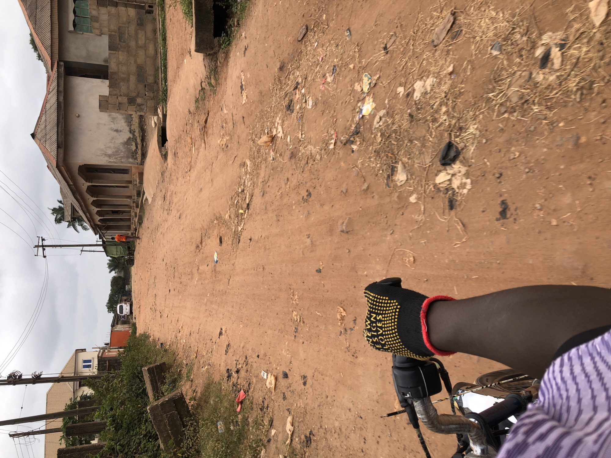 The image captures a street scene in Agege, Nigeria, from the perspective of someone riding a motorcycle. The road is unpaved and covered in red dirt, with visible debris and trash scattered on both sides. On the left, a house with a beige roof and white walls is partially visible, with some overgrown vegetation and concrete pillars. Further down the street, a white van is parked. The right side of the street features a row of buildings with corrugated metal roofs, some with visible brickwork and arched doorways. In the foreground, the rider's hand wearing a black and yellow gloved hand is visible on the handlebars of the motorcycle. The sky is overcast with clouds.