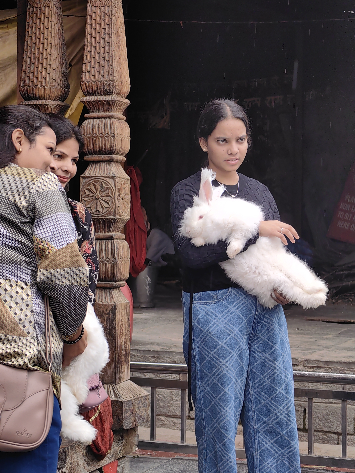 In Vashisht, India, three individuals are positioned near ornately carved wooden pillars. On the left, two women stand close together, with one partially obscured. One of these women is holding a white rabbit, which is also partially visible. To the right, a young woman, also holding a white rabbit, looks towards the left. The rabbit she holds is fluffy with pink eyes and long ears. She is wearing a black long-sleeved shirt and blue patterned pants. The background features a dark structure with some text and what appears to be falling rain. A railing is visible in the foreground, and a light-colored handbag with a strap is slung over the shoulder of the woman on the far left. The overall setting suggests an outdoor environment, possibly a market or temple area in Vashisht.