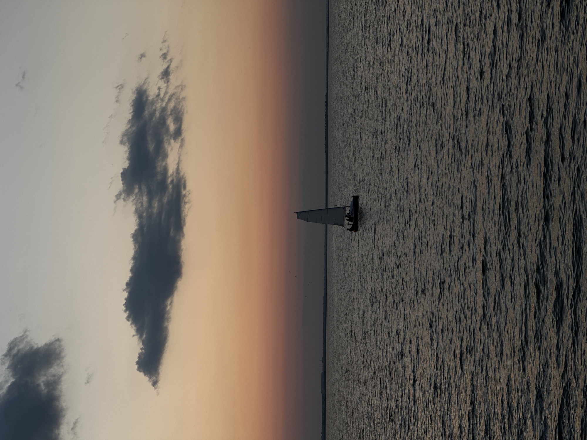 A sailboat with two people onboard is sailing on a body of water during twilight. The sky is a gradient of pale yellow and orange with dark gray clouds. The horizon line is visible, with a distant landmass or city skyline. The water is dark and slightly rippled, reflecting the dim light. The scene is tranquil and serene. The image does not provide any specific indicators of the location in the city of Kokshetau, Kazakhstan.
