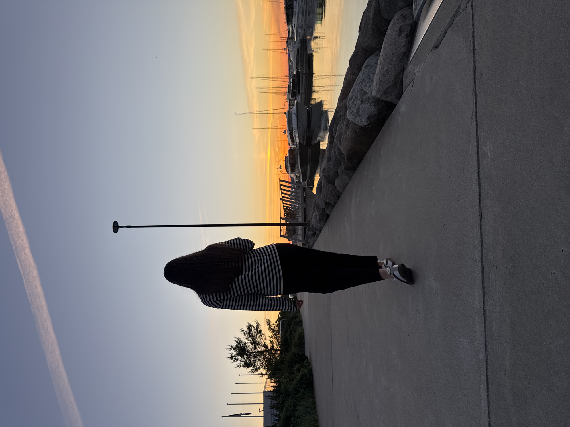 A lone woman with long, dark hair walks away from the camera on a paved walkway. She is wearing a horizontally striped long-sleeved shirt and dark pants, with silver athletic shoes. The walkway runs alongside a harbor filled with sailboats and other watercraft. The sky is a gradient of blue to orange, indicating sunset, and a single contrail is visible in the upper portion of the sky. The harbor water reflects the colorful sky and boats. The walkway is bordered by large rocks on the right and a grassy area with trees and flags on the left. The scene is likely located in a marina in Tallinn, Estonia, possibly Pirita Marina, given the presence of flags and the general ambiance of a seaside city.