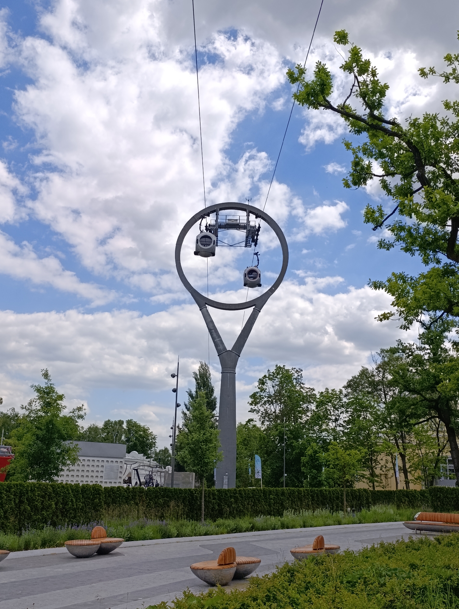 The image showcases a modern cable car pylon standing prominently against a bright, partly cloudy sky. The pylon is a sleek, gray structure with a distinctive Y-shaped base that extends upwards to form a large, open circular frame at its peak. Within this circular mechanism, two small, contemporary gray cable car gondolas are visible, suggesting the system is operational. One gondola hangs near the top, while the other appears to be in motion, passing through the circular pylon.
The setting is an urban park or public space, characterized by abundant greenery. Lush green trees, some with broad leaves and others with more conical shapes, frame the pylon and extend into the background. A manicured dark green hedge runs along a paved pathway in the mid-ground, separating the path from grassy areas dotted with smaller plants and shrubs. Along the pathway, several modern, aesthetically designed benches with wooden slatted seats set into concrete bases are visible, inviting relaxation. In the background, partially obscured by trees and the hedge, urban structures including a light-colored building with a textured pattern and tall streetlights can be seen.
The time of day is daytime, likely mid-day, given the bright and even lighting. The sky is a clear blue, adorned with scattered white cumulus clouds, indicating fair weather. The overall impression is one of a well-maintained, modern public amenity in an urban environment. The location is Moscow, Russia. No people are visible, and there is no visible text.