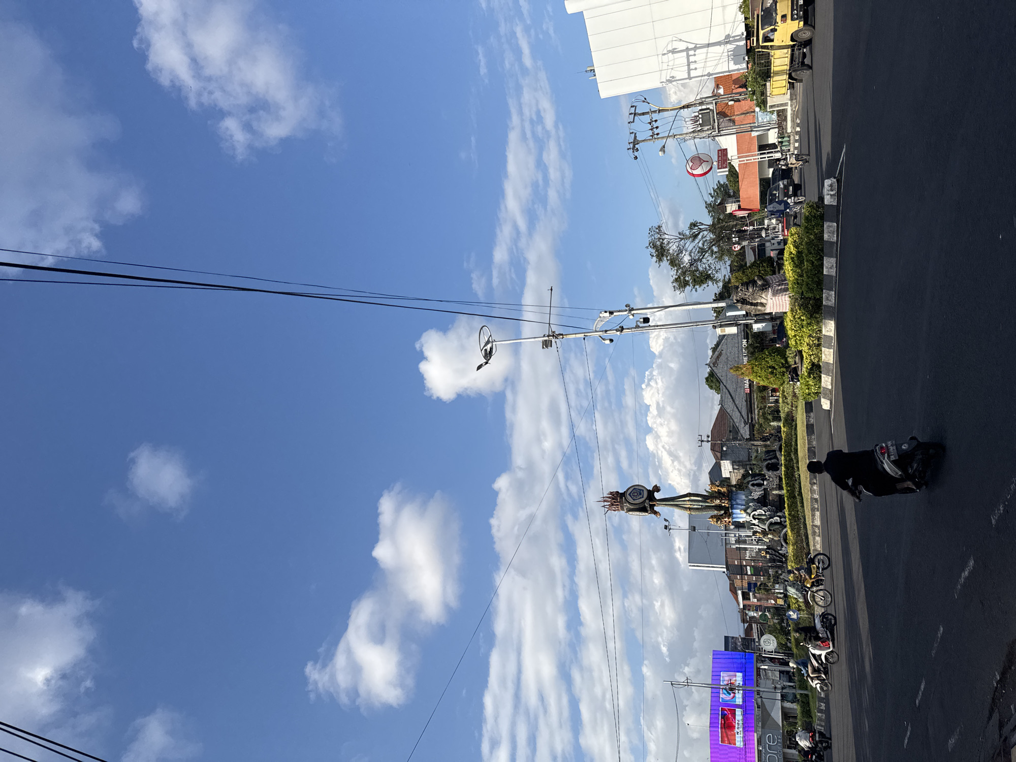 The image shows a view of a busy intersection in Denpasar, Indonesia, with a large monument in the center of a roundabout. The monument features sculptures of mythical creatures and a clock tower. The sky is a clear blue with scattered white clouds, and power lines crisscross the frame. Several motorcycles are visible on the road, with riders engaged in various activities. One person on a scooter is facing away from the camera, while others are moving in different directions. In the background, buildings of various sizes and architectural styles line the street, including a large, modern white building on the right and a digital billboard on the left displaying advertisements. A yellow truck is also present in the background. Lush green bushes and trees surround the roundabout, adding a touch of nature to the urban landscape. The overall scene depicts a vibrant and active street in Denpasar.