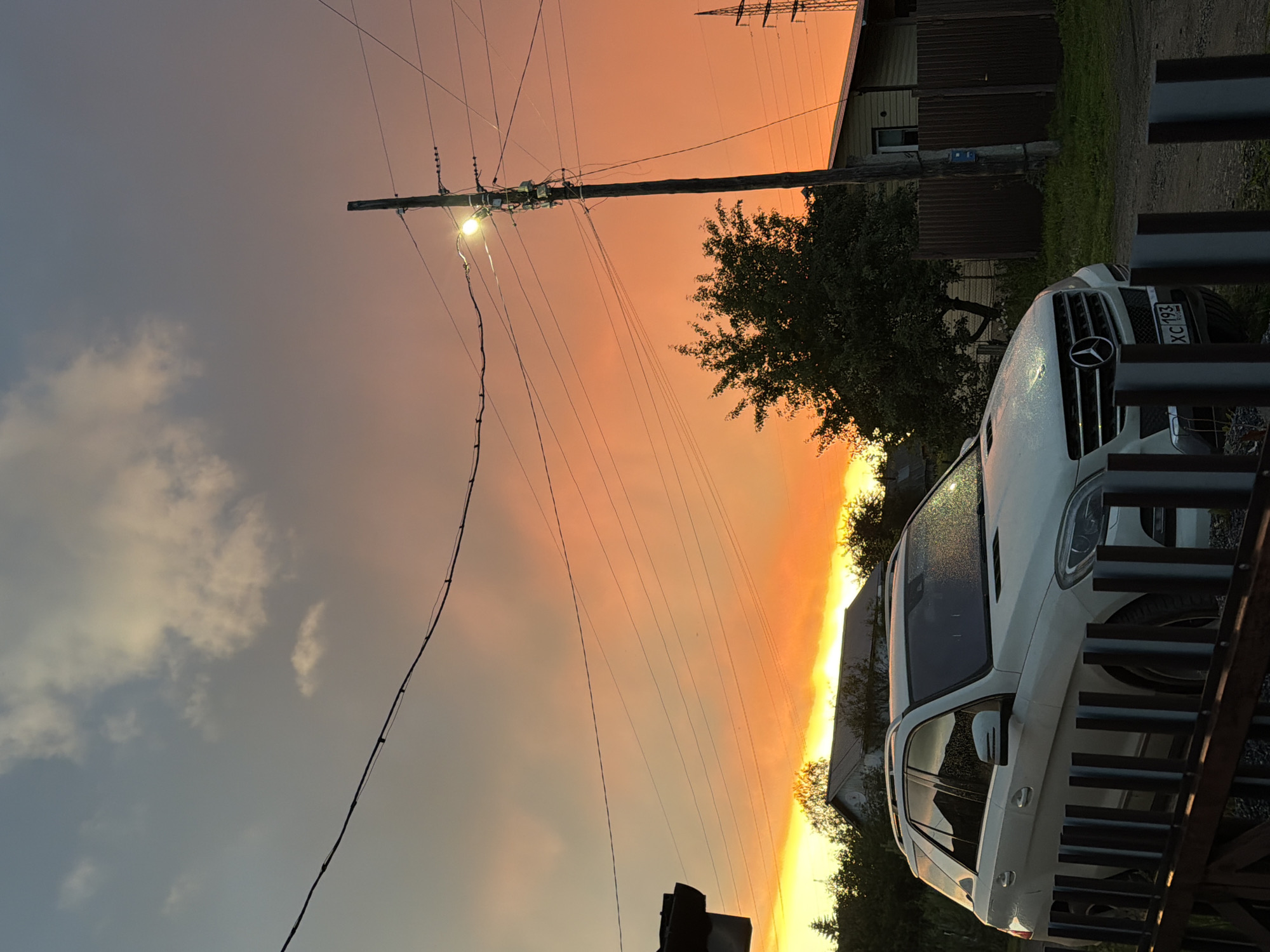 This vertical-format media file depicts a sunset scene in Mezhevoy, Russia. The sky is dominated by a gradient of colors, transitioning from a dusky blue on the left to vibrant orange and yellow hues on the right, indicating it's likely late evening. Scattered clouds are visible in the bluer portion of the sky.
Several man-made elements are present. A wooden utility pole with multiple wires crisscrossing it stands prominently in the middle of the frame, with a lit bulb hanging from it. In the background, a building with a corrugated metal fence and some trees are visible.
In the foreground, a white Mercedes-Benz SUV is parked. The vehicle's paint appears slightly wet, possibly from recent rain, and reflects the colors of the sunset. The license plate is partially visible and reads "XC 493". The overall atmosphere conveyed is one of a peaceful, rural evening.
