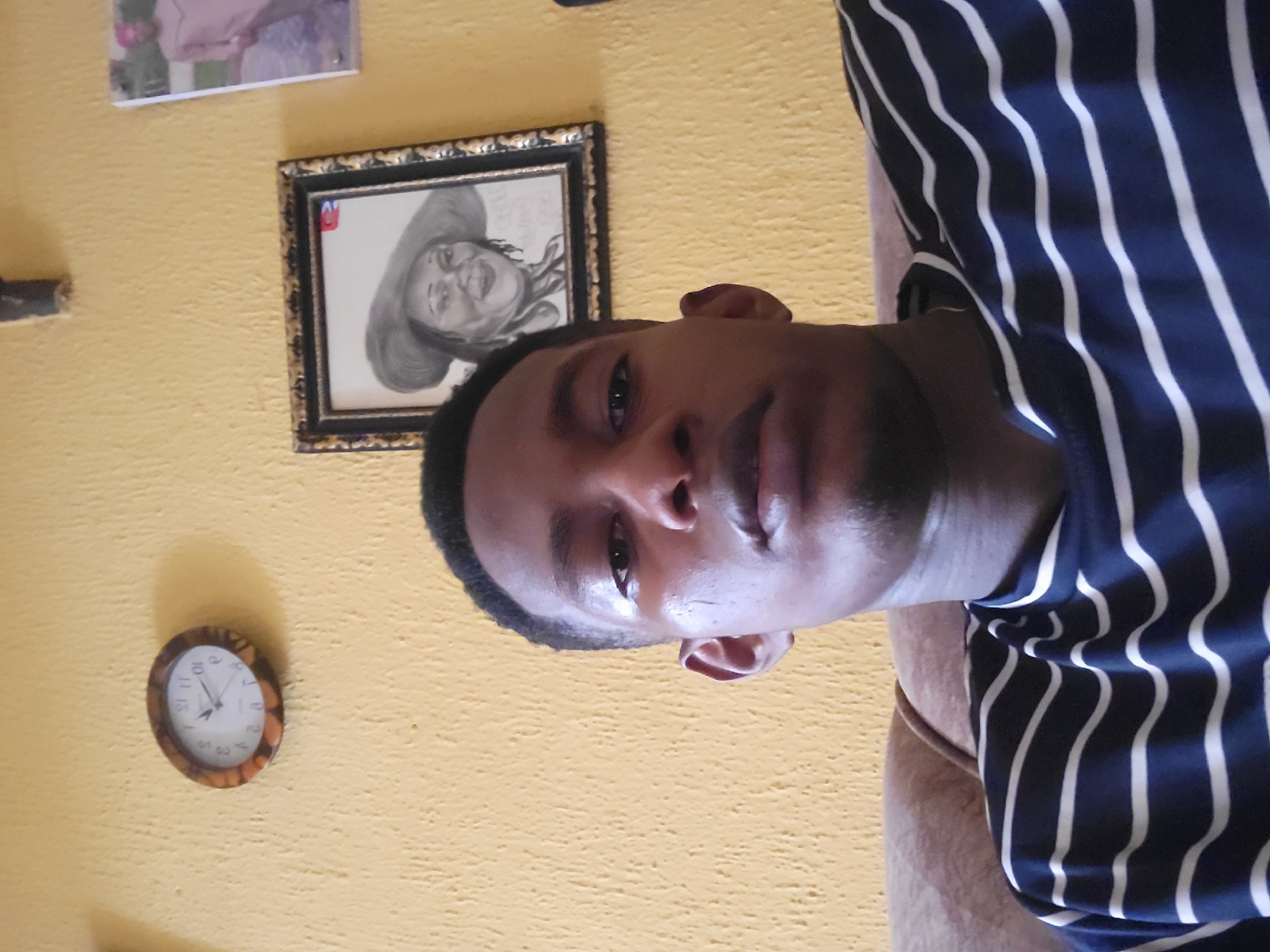 The image is a selfie taken by a young man in his home. The man is wearing a striped t-shirt and is seated on a couch. In the background, a yellow, textured wall is visible. On the wall, there are two framed pictures and a clock. One picture is a sketch of a woman wearing a hat, and the other is a photograph of a person in traditional Nigerian attire. A circular clock with Roman numerals is also on the wall. The scene appears to be a casual indoor setting, likely a living room. It is not possible to determine the exact location within Onitsha, Nigeria, based solely on this image.