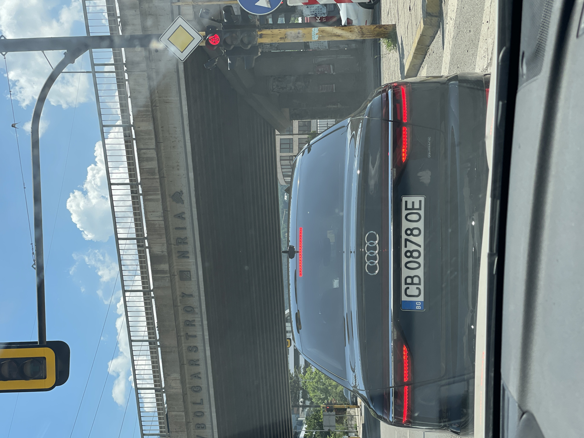 The image shows the rear of a dark-colored Audi sedan stopped at a traffic light in Sofia, Bulgaria. The car has a Bulgarian license plate that reads "CB 0878 OE". The traffic light is red with an arrow pointing upwards, indicating that traffic should proceed straight. Above the car, a concrete bridge crosses the road, with the word "BOLGARSTROY" visible on its side. The sky is blue with some clouds. The scene appears to be a typical urban environment with traffic, infrastructure, and signs.
