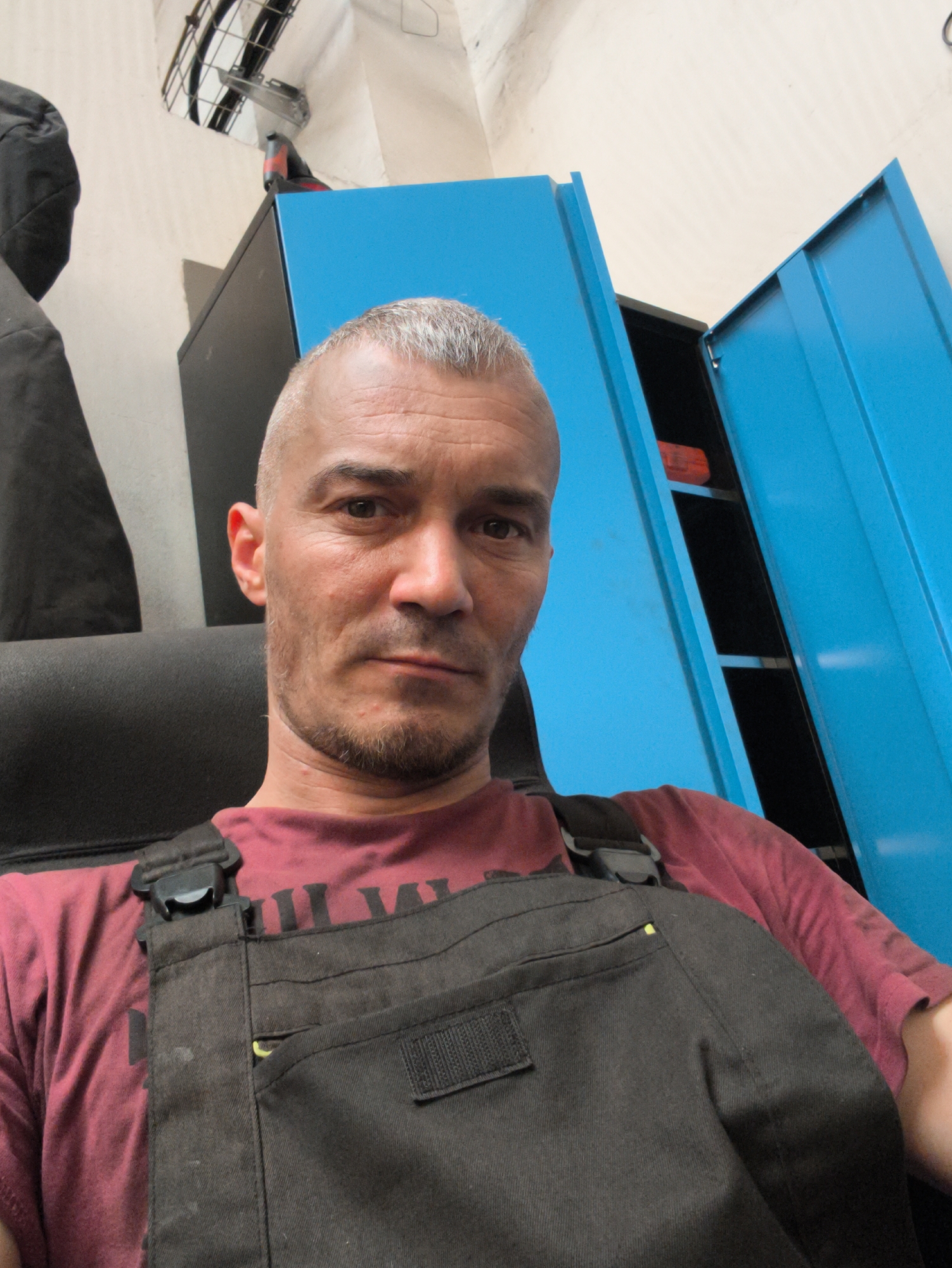 This is a selfie of a man with short grey hair and a light beard, wearing a maroon t-shirt and dark grey overalls. He is sitting in a black chair, with his upper body and face filling most of the frame. Behind him, there are bright blue metal cabinets with one door open, revealing black shelves and a red object. To the left, a dark coat hangs on a wall. The background suggests an indoor setting, possibly a workshop or garage, with fluorescent lighting visible overhead. The man's expression is neutral. The time of day and weather are not discernible. There is no visible text in the image.