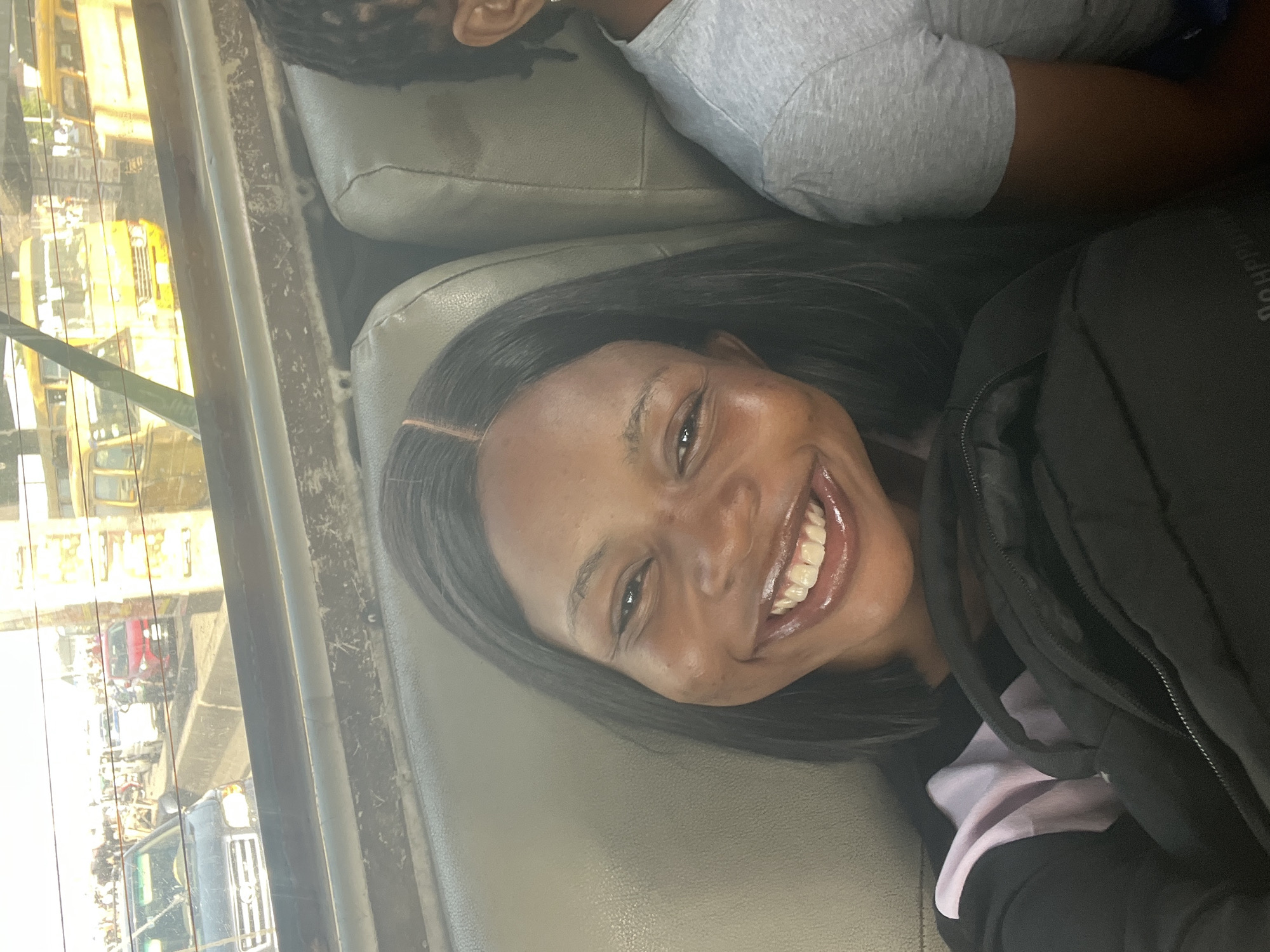 The image captures a cheerful moment inside a vehicle, likely in Satellite Town, Nigeria. The primary subject is a young woman, positioned in the foreground, smiling broadly and looking directly at the camera. She has long, dark hair parted in the middle and dark skin. She is wearing a black jacket or hoodie over a light-colored garment, with a black bag or backpack partially visible next to her that features indistinct, partially visible text.
Behind her, another person's head (top and ear) and a grey t-shirt are partially visible, suggesting they are seated in the vehicle's row behind. The vehicle's interior features grey or tan upholstered seats.
Through the rear window, a busy daytime street scene unfolds. Bright sunlight illuminates various vehicles, including several distinct yellow buses or large commercial vehicles, a red car, and other traffic. Buildings and structures line the street, indicating an urban or semi-urban environment. The overall impression is one of happiness during travel or transit on a sunny day.
