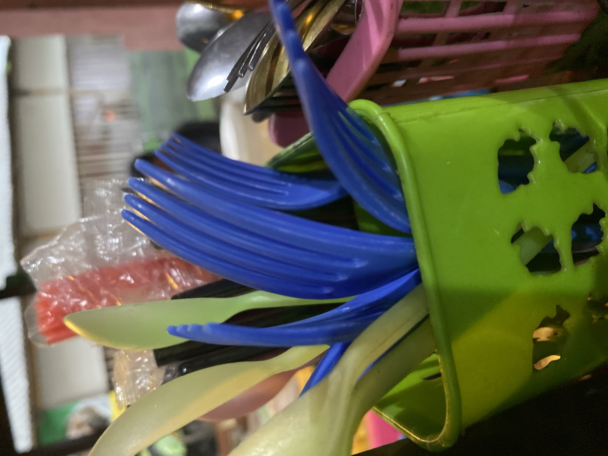 The image displays a close-up view of a collection of plastic cutlery, predominantly forks and spoons, stored in a green plastic holder. The cutlery is a mix of colors including blue, light yellow, and some black. In the background, a pink plastic basket is visible, also containing cutlery, along with other items that are out of focus, suggesting an outdoor setting or a space with ample light. The overall impression is one of everyday, casual dining or preparation. The location is identified as Kasihan, Indonesia, which is a subdistrict in the Bantul Regency, Yogyakarta Special Region. While the specific context within Kasihan is not evident from the image itself, the scene suggests a place where food service is common, possibly a street food stall or a casual eatery.