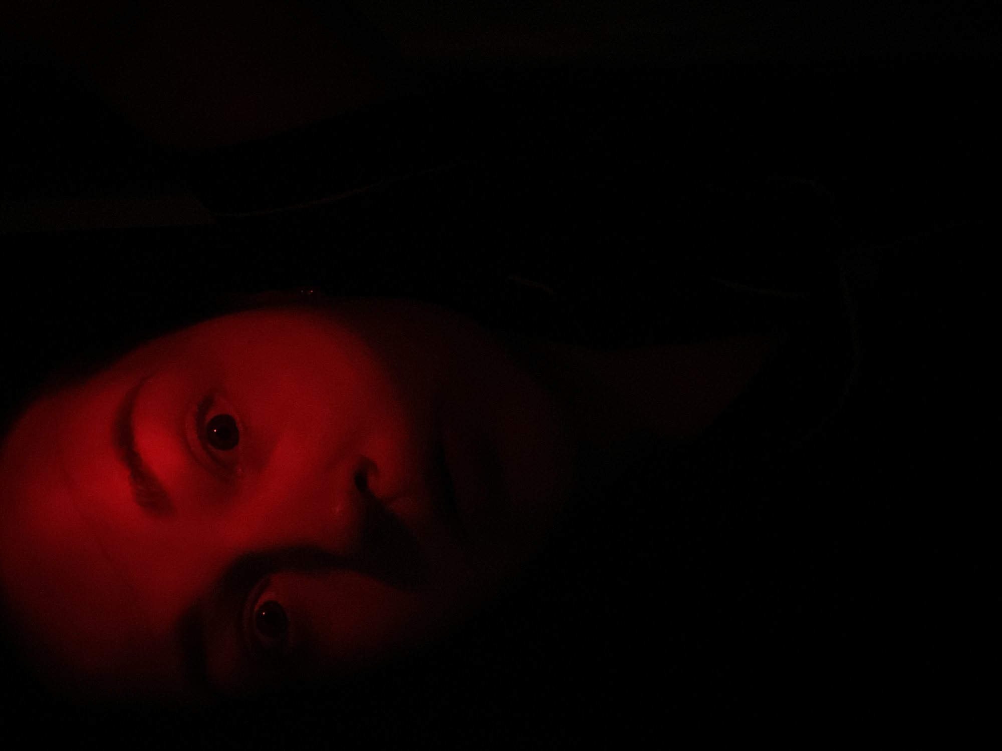 The image presents a close-up of a person's face, dramatically illuminated by an intense red light against a completely dark background. The face is positioned on the left side of the frame, tilted, with the top of the head towards the upper left. The subject's eyes are wide open, their pupils visible and reflecting the red light, creating a focal point. Visible features include the eyebrows, the bridge and tip of the nose, and a portion of the mouth and chin, all rendered in deep red tones and contrasting shadows. The dominant red lighting creates a stark, high-contrast effect, emphasizing the facial contours and giving the scene a mysterious or intense emotional quality.
The setting appears to be extremely dark, suggesting either nighttime or an enclosed, unlit space. There are no other objects or discernible elements in the black background. The activity shown is the subject gazing directly forward, possibly at the viewer or an unseen light source. There are no visible location cues within the image itself to indicate the provided context of Sochi, Russia. No text is visible.
