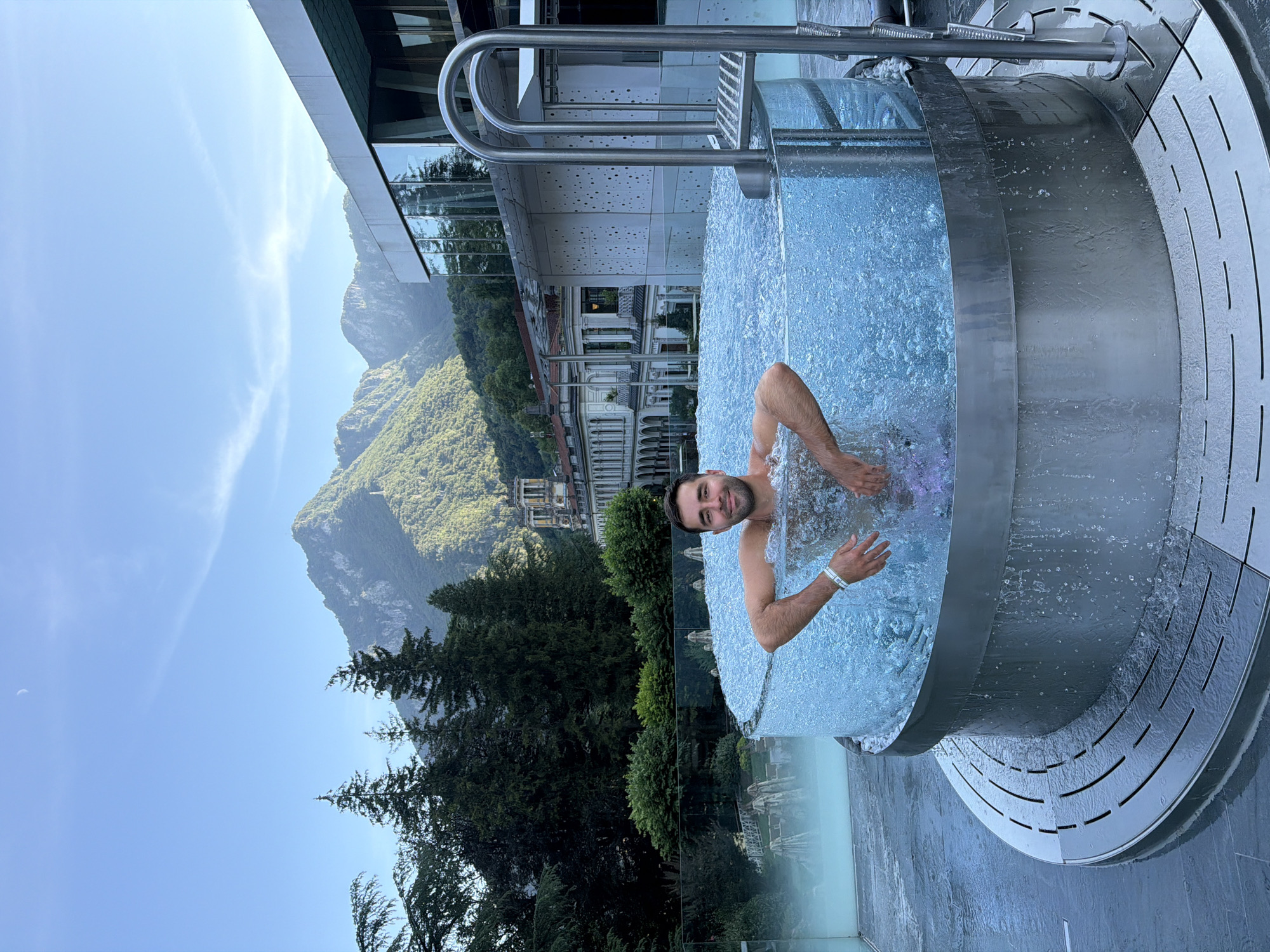 In San Pellegrino Terme, Italy, a man relaxes in a clear-walled, circular hot tub filled with bubbling water. The hot tub is situated on a tiled patio with a metal grate around its base. A metal ladder with two steps and a handrail is attached to the side of the tub. In the background, a modern building with large glass windows stands adjacent to older, traditional Italian architecture. Beyond the buildings, lush green mountains rise under a clear blue sky with a crescent moon visible. Trees line the landscape between the hot tub and the buildings. The man, with dark hair and a beard, is smiling at the camera, his arms resting on the edge of the tub. Water splashes around him.