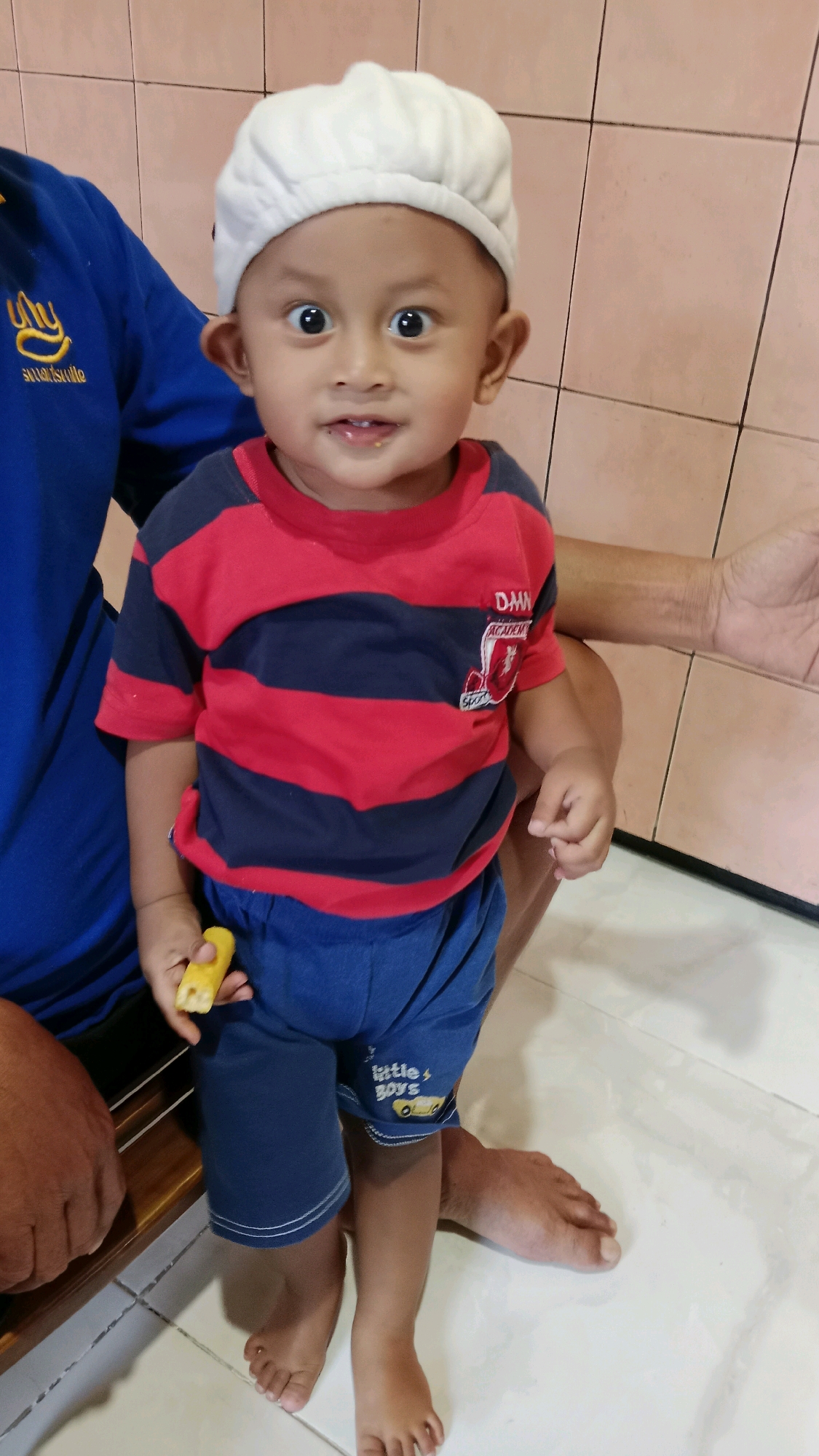 In a scene indoors, likely in Kebonagung, Indonesia, a young child, approximately two years old, stands and holds a piece of food in their right hand. The child is wearing a white beret, a red and dark blue striped t-shirt with a crest on the left chest, and dark blue shorts with white trim and a graphic. Their large eyes are open and directed towards the viewer, and their mouth is slightly open. To the left of the child, a person in a blue shirt is partially visible. To the right and behind the child, another person's arm and hand are visible. The background consists of tiled walls and a tiled floor.