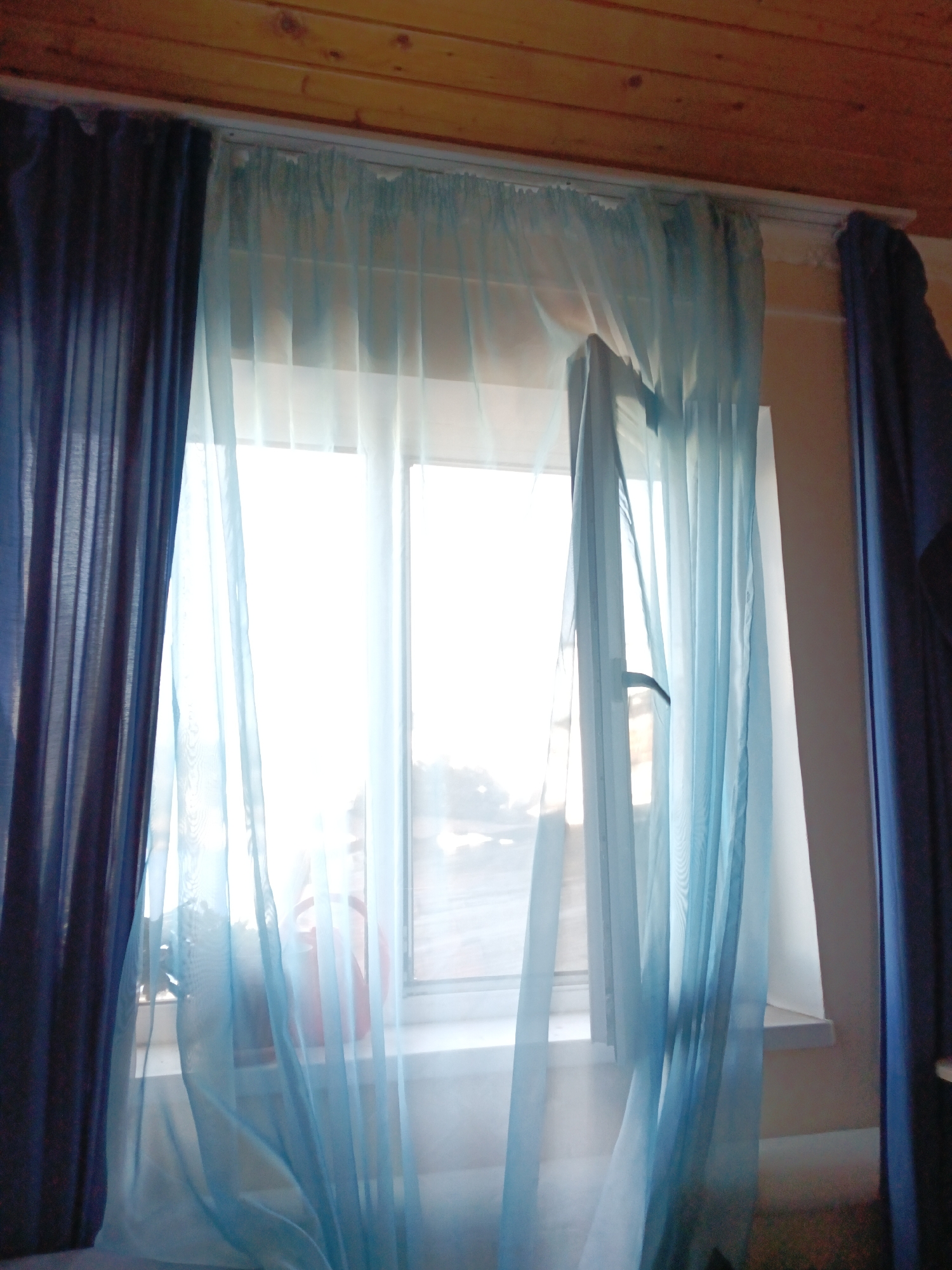 A medium shot captures a window framed by sheer blue curtains and darker blue drapes. The window itself is illuminated from the outside, creating a bright, hazy effect. A light blue sheer curtain is drawn across the window, with a section of it appearing to be opened or pulled aside, revealing a partially obscured view. On the windowsill, a pale pink watering can is visible, with some indistinct shapes behind it. The ceiling above is made of wooden planks, and the wall to the right of the window is a plain beige. The scene appears to be indoors, likely in a residential setting. No people are visible in the image.