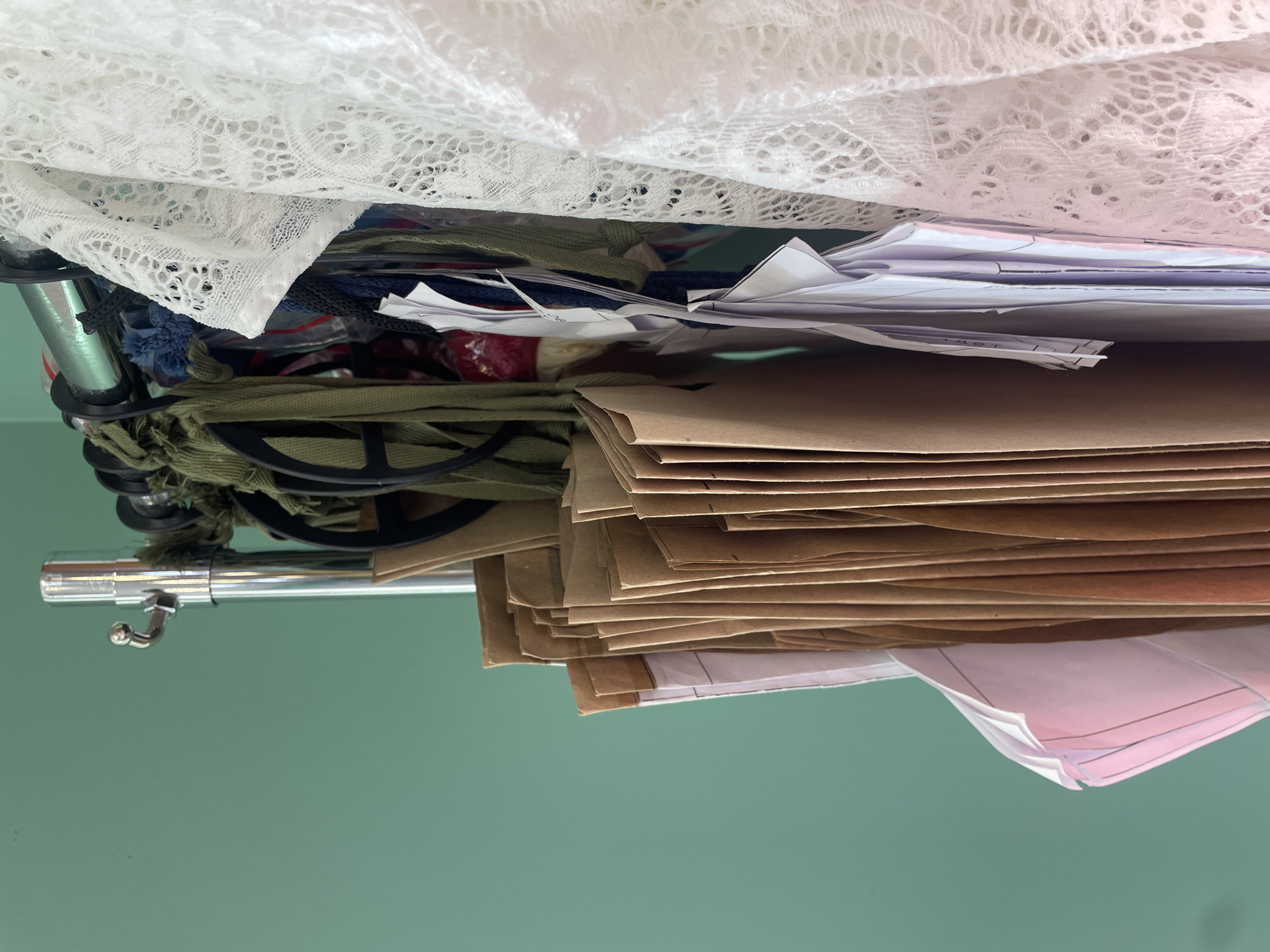 A close-up, eye-level shot shows a rack of sewing patterns and a white lace garment against a teal-colored wall. The sewing patterns are made of brown paper and white paper, and they are folded and hanging on the rack. The white lace garment is draped over the rack, and it is a close-up of its texture. The background is a solid teal color. The image is lit from the left, casting shadows on the right side of the objects.