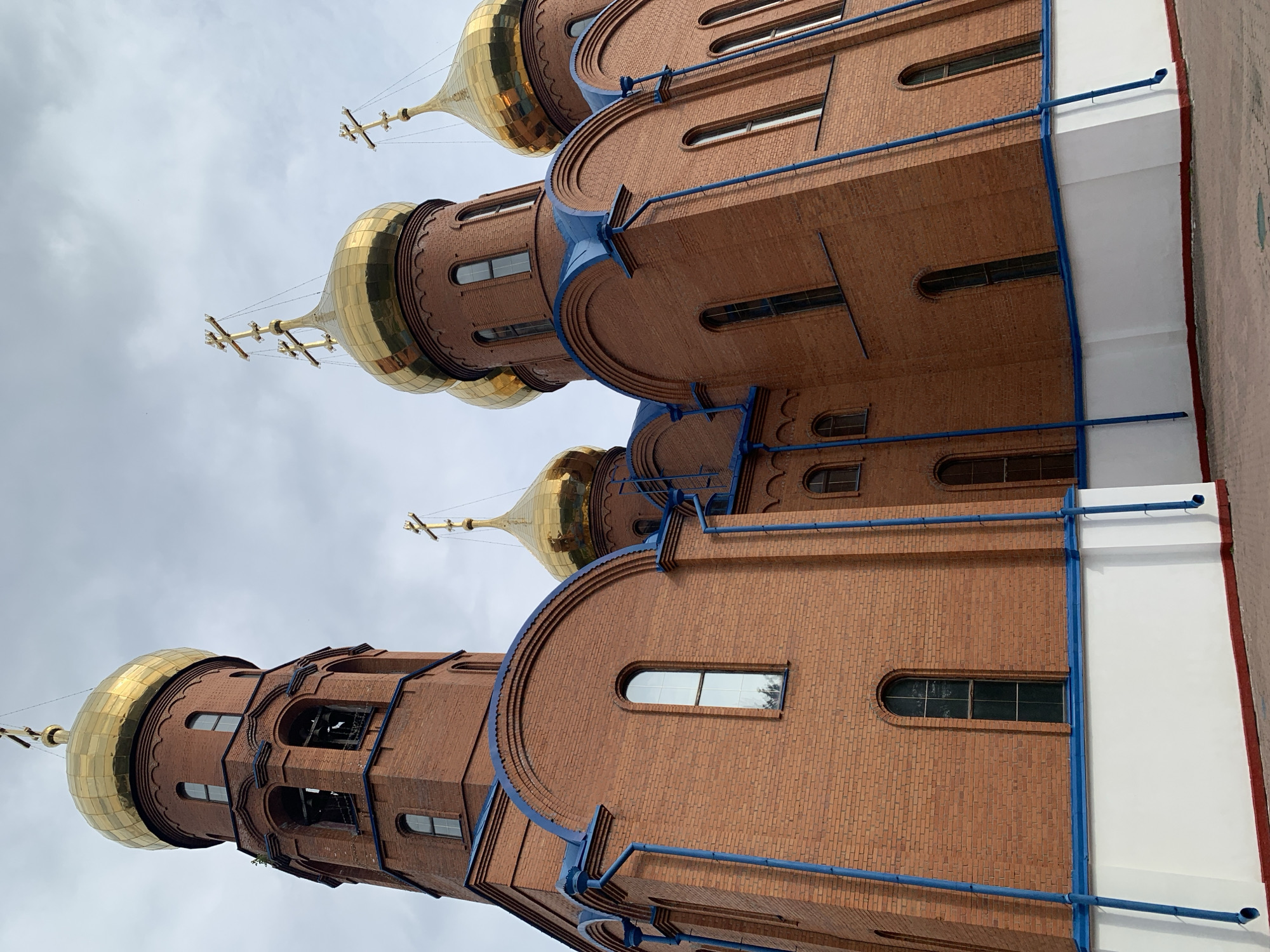 The image shows a close-up, low-angle shot of the brick exterior of an Orthodox church in Prokopyevsk, Russia. The church features multiple golden domes topped with crosses, and blue trim along the rooflines and downspouts. The brickwork is a reddish-brown color, and the building has arched windows. The sky is overcast. The scene is devoid of people and any discernible action. The ground in the foreground is paved.