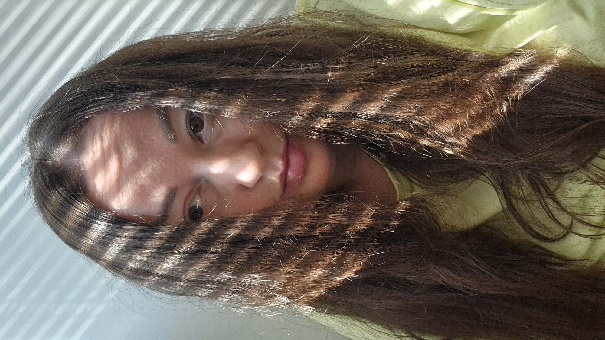 This media file presents an intimate, close-up portrait of a young woman, likely taken indoors in Kemerovo, Russia. Her long, dark brown hair, with natural highlights, dominates the frame, flowing around her face and over her shoulders. The scene is illuminated by strong natural sunlight, which casts distinct, parallel striped shadows, presumably from venetian blinds, across her forehead, the bridge of her nose, and various sections of her hair, creating a captivating interplay of light and shadow.
The woman looks directly at the camera with her dark eyes. Her mouth is slightly open, revealing a hint of her tongue, lending a playful or relaxed quality to her expression. Her skin appears naturally textured and well-lit by the sun. She is wearing a light-colored garment, possibly pale green or yellow, visible on the right side of the image beneath her hair. The background behind her head is a light, unidentifiable surface, also marked by the striped light patterns. The clear, directional sunlight suggests it is daytime with sunny weather. The overall impression is one of a candid and natural moment, possibly a self-portrait. No visible text is present in the image.