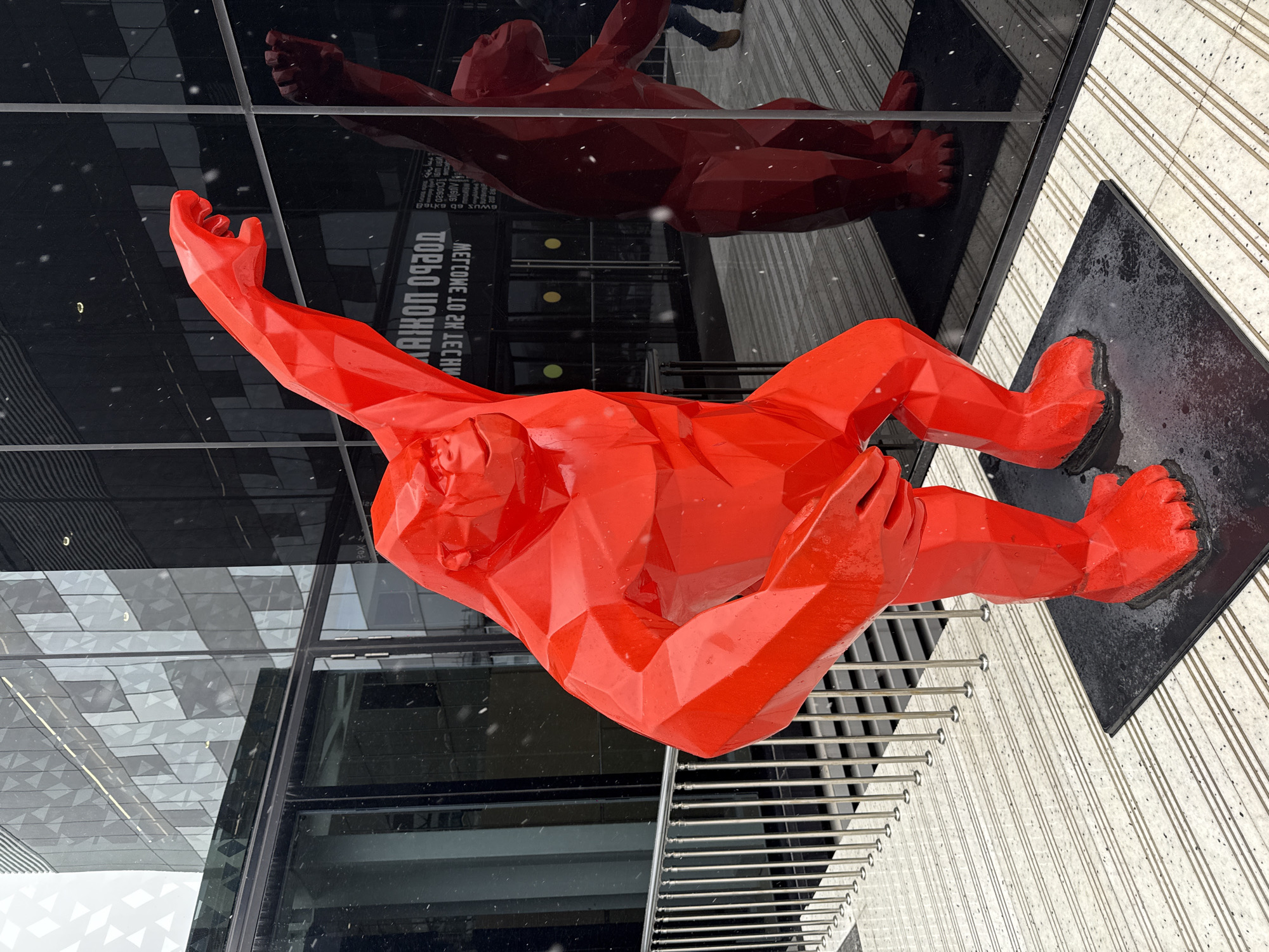 The image displays a large, vibrant red, faceted sculpture of a human figure positioned horizontally outdoors on a snowy day. The sculpture, rendered in a low-poly style, depicts a reclining or falling male figure with outstretched limbs; one arm is raised and open-palmed, while a leg is bent. It rests on a dark, rectangular base.
Behind the sculpture stands a modern building with a dark, highly reflective glass facade. The glass perfectly mirrors the red sculpture as a darker, inverted image, along with other architectural details and the legs of a person standing nearby. The reflection also clearly shows inverted and reversed Russian text on the building's entrance. Visible phrases include "ДОБРО ПОЖАЛОВАТЬ" (Welcome), "МЕТРОМ 10 МИНУТ" (10 minutes by metro), and "ТЕЛЕФОН:" (Telephone:) followed by numbers.
The ground features light-colored, textured paving with parallel dark lines, and a metal grate structure is visible to the left of the sculpture. Small white snowflakes are scattered throughout the scene, indicating a cold, wintery day. The overall setting is a modern urban environment in Moscow, Russia, where contemporary art interacts with sleek architecture under snowy conditions during daylight hours.