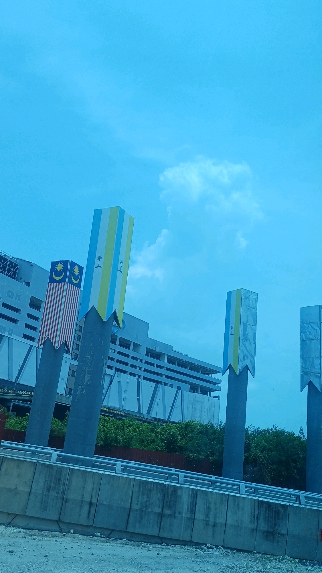 The image captures a scene in Butterworth, Malaysia, featuring several large, decorative pillars with various designs. One pillar on the left displays the Malaysian flag. Another pillar to its right is adorned with vertical stripes of blue, yellow, and white, each featuring a palm tree motif. The two pillars further to the right have a similar stripe pattern but appear to be covered in a metallic material.
Behind these pillars, a multi-story building, possibly a parking structure or commercial building, is visible. The building has a modern architectural style with a light-colored facade and numerous windows or openings. Lush green vegetation lines the area below the building, creating a contrast with the concrete structures.
The foreground is dominated by a concrete barrier, likely part of an elevated road or highway, with metal railings along the top. The ground in the immediate foreground appears to be a mix of dirt and small stones. The sky above is a bright blue with scattered white clouds, suggesting daytime. The overall impression is an urban landscape with public art installations.