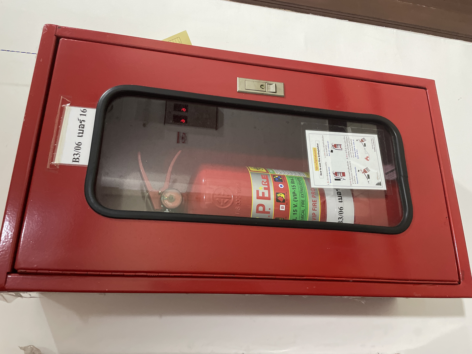 A bright red metal fire extinguisher cabinet is mounted on a white wall. The cabinet has a glass door with a black rubber seal, through which a red fire extinguisher is visible. A label on the top right of the cabinet reads "B3/06 เบอร์ 16". Inside the cabinet, above the extinguisher, are two red digital displays, each showing the digit "a". A silver latch with a "PUSH" button is located on the right side of the cabinet door. A white instructional diagram titled "How to use Fire Extinguisher" is affixed to the inside of the cabinet door, partially obscuring the fire extinguisher. The environment suggests an indoor location, likely a public building or facility within Pattaya City, Thailand, given the presence of Thai script on the label.