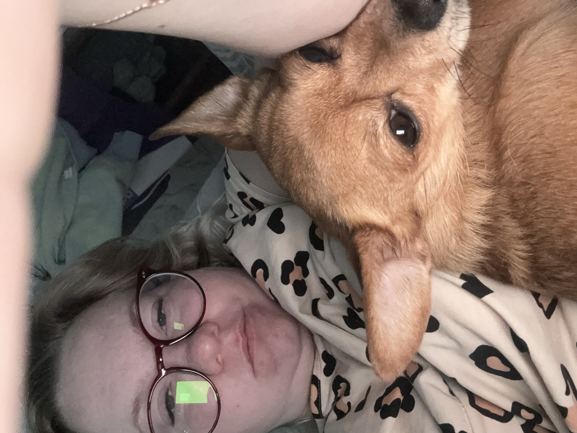 A selfie taken from a slightly elevated angle shows a woman with blonde hair and round glasses lying down next to a tan-colored dog. The woman is facing upwards towards the camera. The dog is positioned next to the woman, with its head resting on her. They are both lying on a surface with a black and tan heart pattern. The location is unknown.