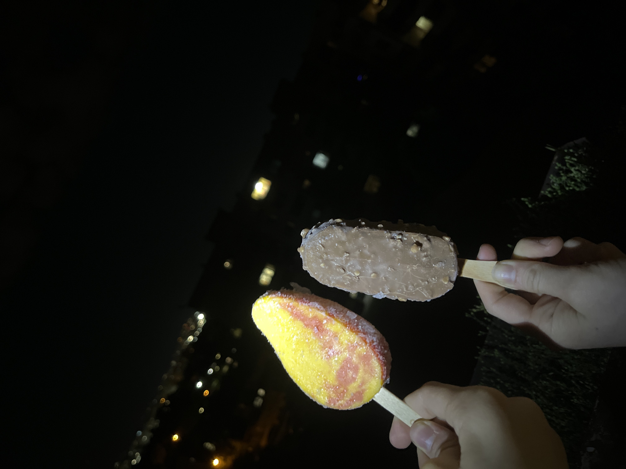 Two hands are holding ice cream bars on sticks, against a dark background. The ice cream in the lower hand is yellow and pink, while the ice cream in the upper hand is brown. Bokeh from a building is visible in the background. Based on the architecture and urban layout, it is plausible that the image was taken in Sofia, Bulgaria.