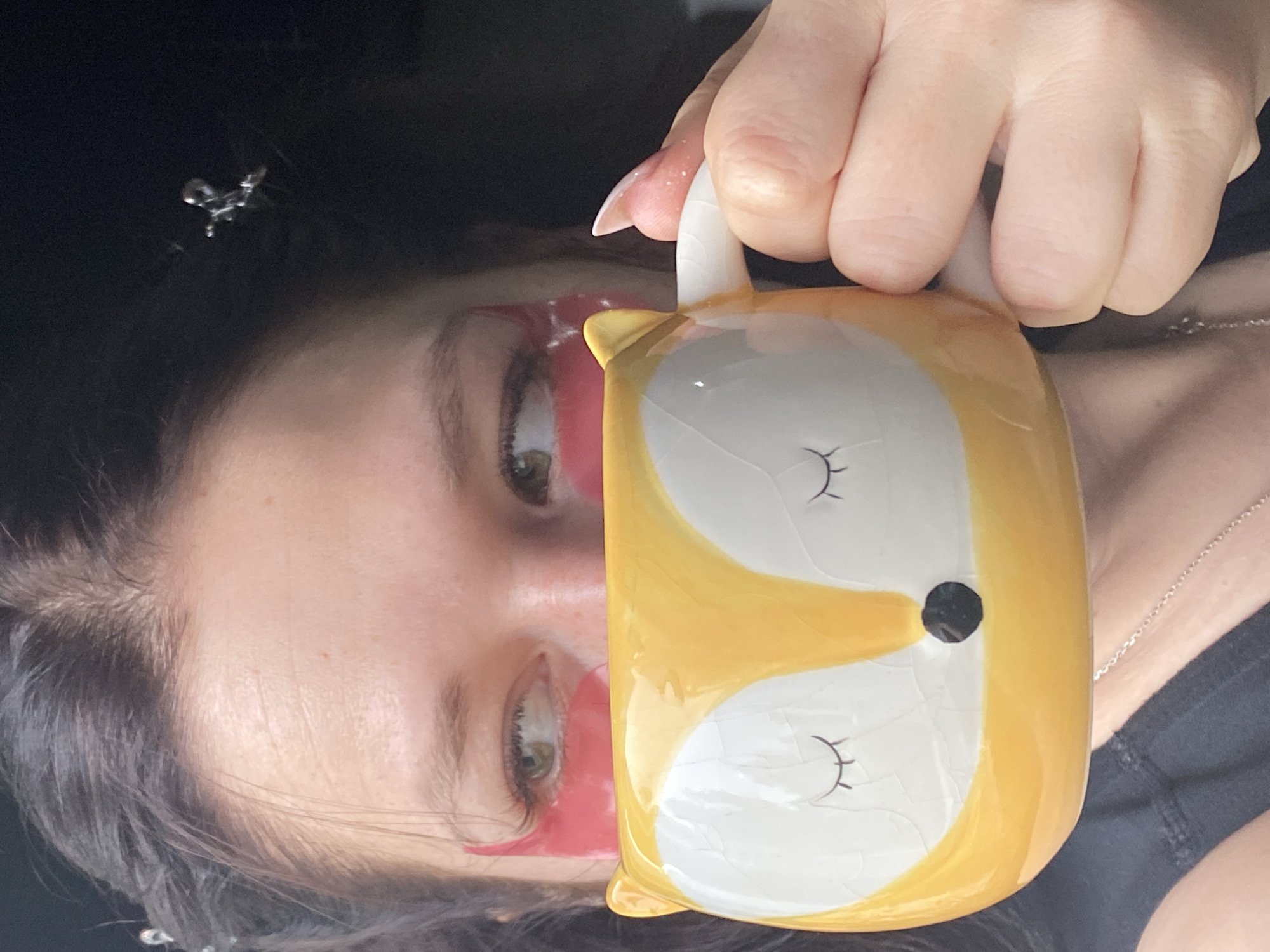A person is holding a yellow and white fox-shaped mug. The person has dark hair and is wearing pink under-eye patches. The person's face is partially visible, with green eyes visible. A silver necklace is visible. The background is dark. The location is unspecified beyond the image.