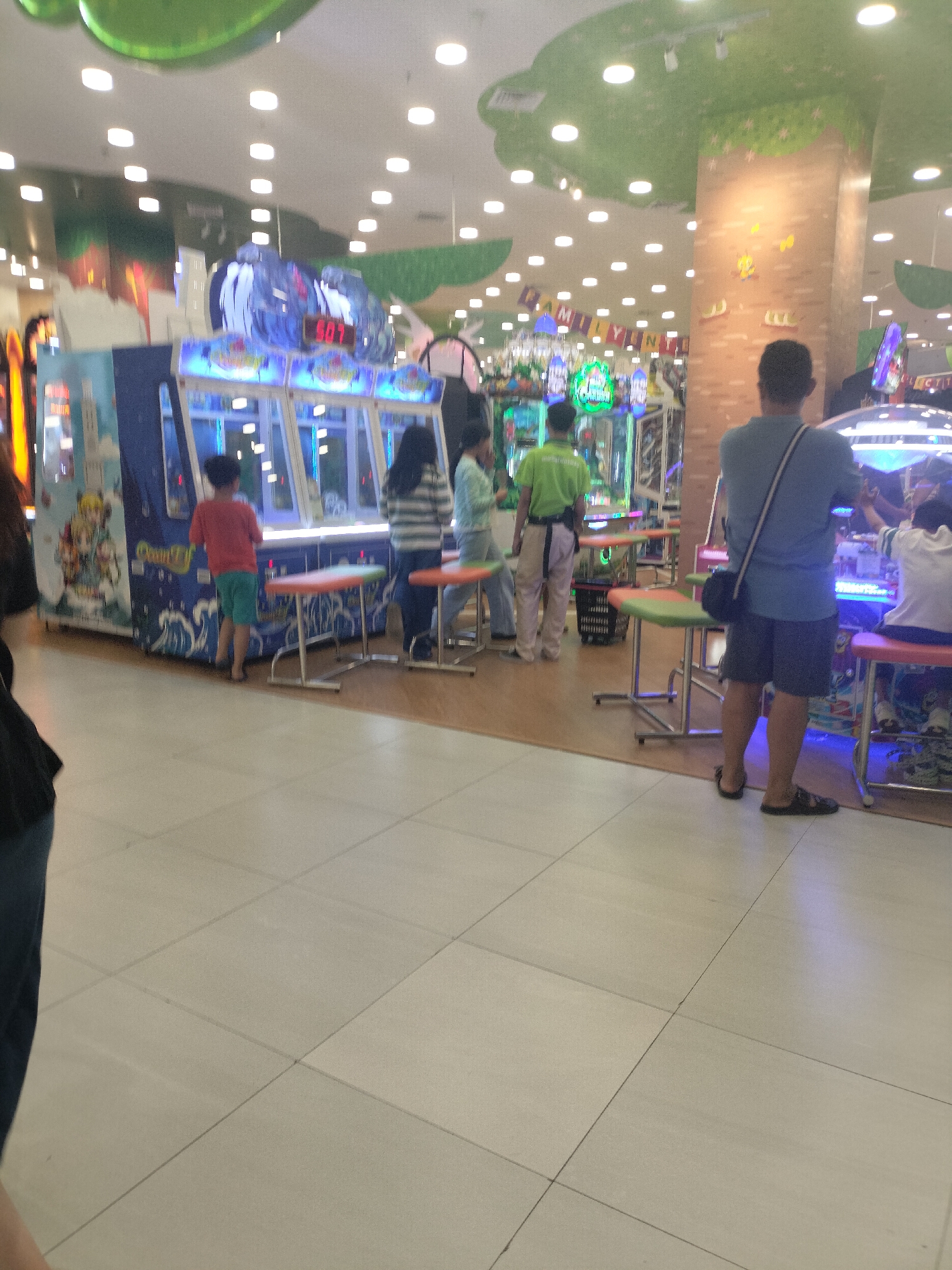The image depicts an indoor arcade or game center, likely located in the city of Gunung Putri, Indonesia. The scene is brightly lit by numerous ceiling lights and illuminated arcade machines. In the foreground, a man in a blue t-shirt and dark shorts stands facing away from the camera, looking towards an arcade game. He has a dark bag slung across his shoulder. To his left, several arcade machines are visible, some with cartoon-like graphics and flashing lights. A young boy in a red t-shirt and green shorts stands in front of one of these machines. To his right, three other individuals are present, two women and one man, all appearing to be engaged with the arcade games. The floor is tiled with light-colored square tiles. In the background, the ceiling has a decorative, possibly green, undulating design, and several large pillars are visible. The overall atmosphere suggests a recreational environment for families or individuals.