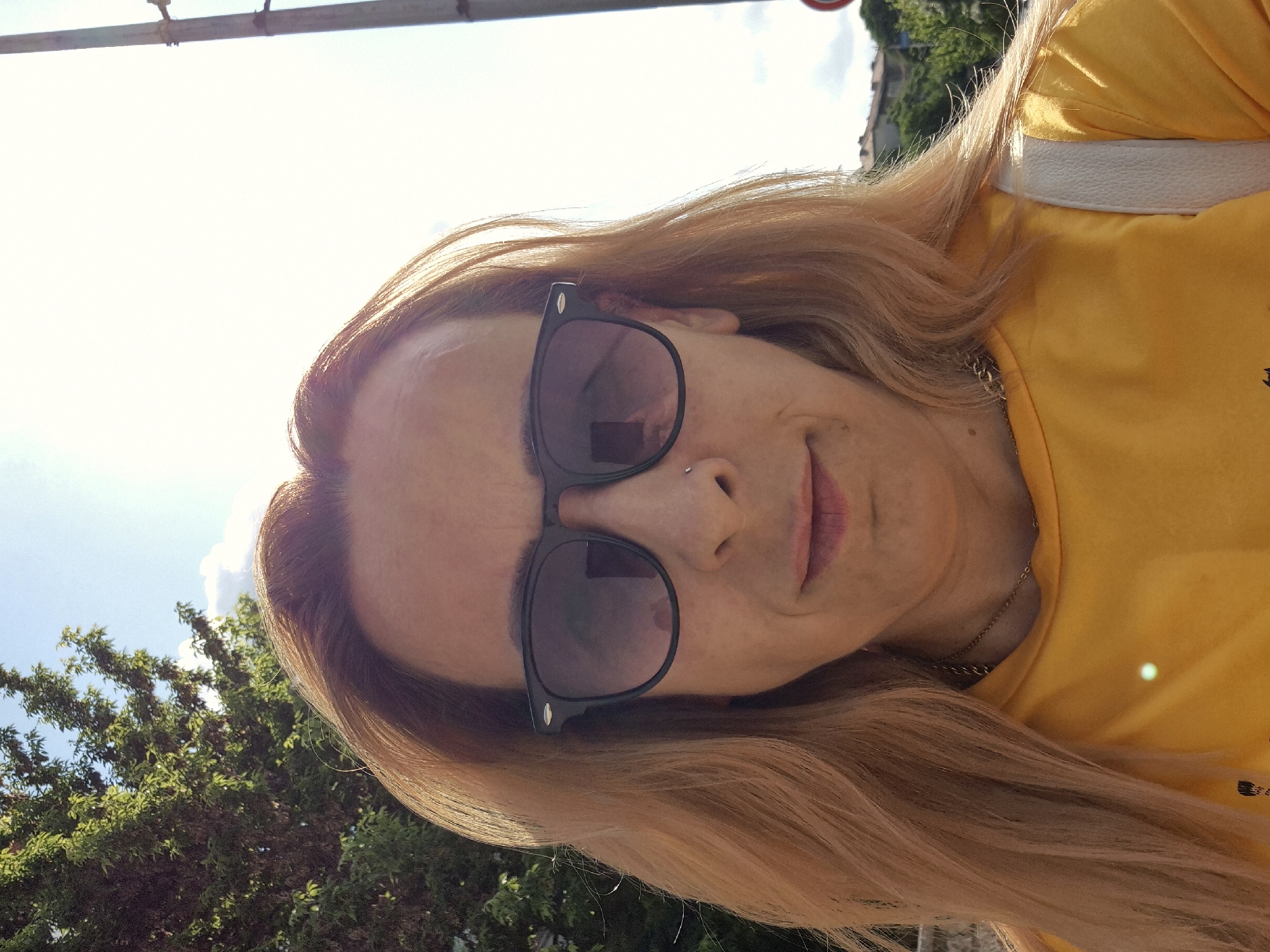This is a close-up, rotated selfie featuring an individual outdoors on a bright, sunny day. The person, with long, flowing light blonde or golden-brown hair, is wearing black sunglasses and a vibrant yellow t-shirt. A thin gold chain necklace is visible around their neck, and they have a small nose piercing. Their expression is subtle, with a slight smile hinted at.
The background consists primarily of a very bright, almost overexposed sky, indicating strong sunlight. Green foliage from trees or bushes is visible in the lower left and middle portions of the background. A metallic bar or structure runs across the top edge of the frame. A white strap, likely from a bag, is visible over the person's right shoulder. A distinct, bright green light reflection appears on the yellow shirt. The overall scene suggests a pleasant daytime outdoor setting. There is no visible text in the image.