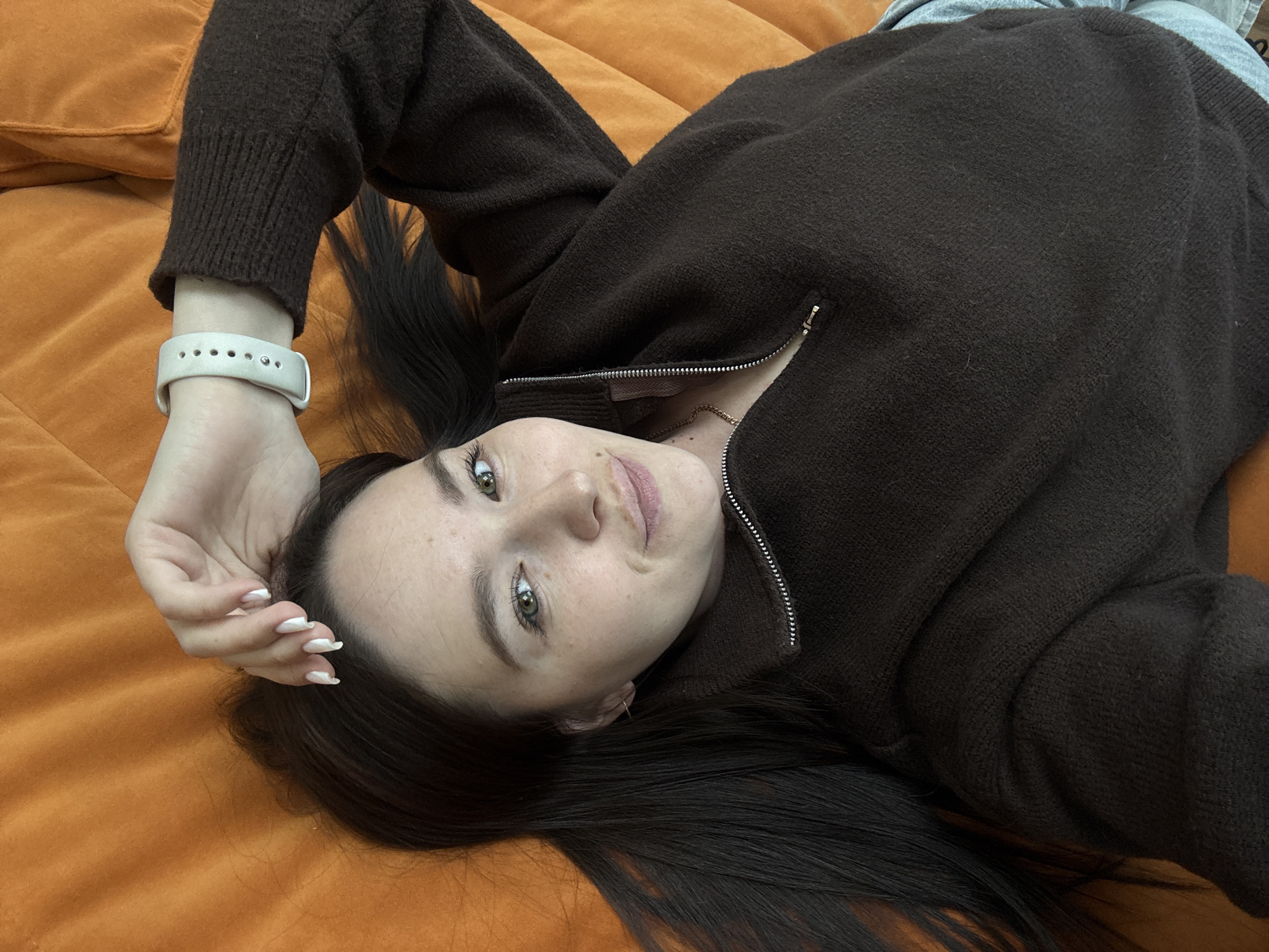A female individual is depicted in a high-angle shot, lying supine on a textured, plain orange surface, consistent with upholstery. Her head is angled towards the upper left, with long dark brown hair fanned out around her. She has light skin with visible freckles, green eyes, and pink-tinted lips, displaying a slight smile while looking directly at the viewer. She is dressed in a dark brown quarter-zip sweater, and a light blue garment is partially visible on her lower right. A thin gold chain is present around her neck. Her left wrist wears a light-colored smartwatch with a perforated band. Her right hand, featuring white, pointed fingernails, is positioned on her forehead, partially obscuring her hairline. The specific location within the city of Chaikovsky, Russia, cannot be determined from the visual information provided.