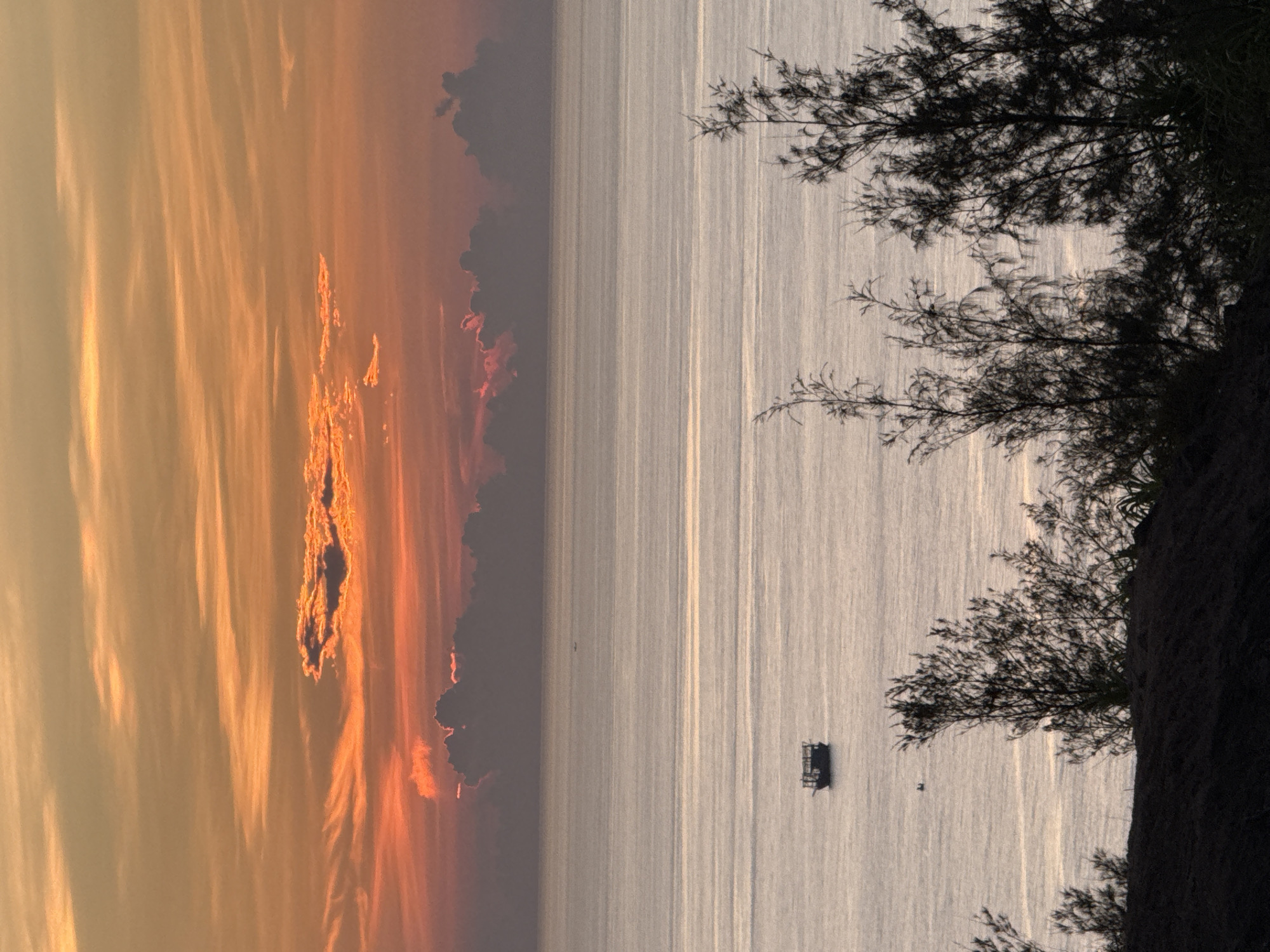 A serene ocean sunset is captured in Lý Sơn Special Administrative Region, Vietnam. The sky is a canvas of warm hues, transitioning from soft oranges and pinks at the horizon to a gentle yellow at the top, with thin, wispy clouds scattered across. A layer of darker, thicker clouds sits low on the horizon, partially obscuring the distant land. The sea is calm, reflecting the sky's colors in subtle, shimmering ripples. In the middle ground, a small, dark boat with a simple structure floats on the water. Closer to the foreground, silhouetted against the dusky light, is a dark mound of land covered with sparse, leafless trees, their branches reaching towards the sky like skeletal fingers. The overall atmosphere is peaceful and tranquil, suggesting the end of a day.
