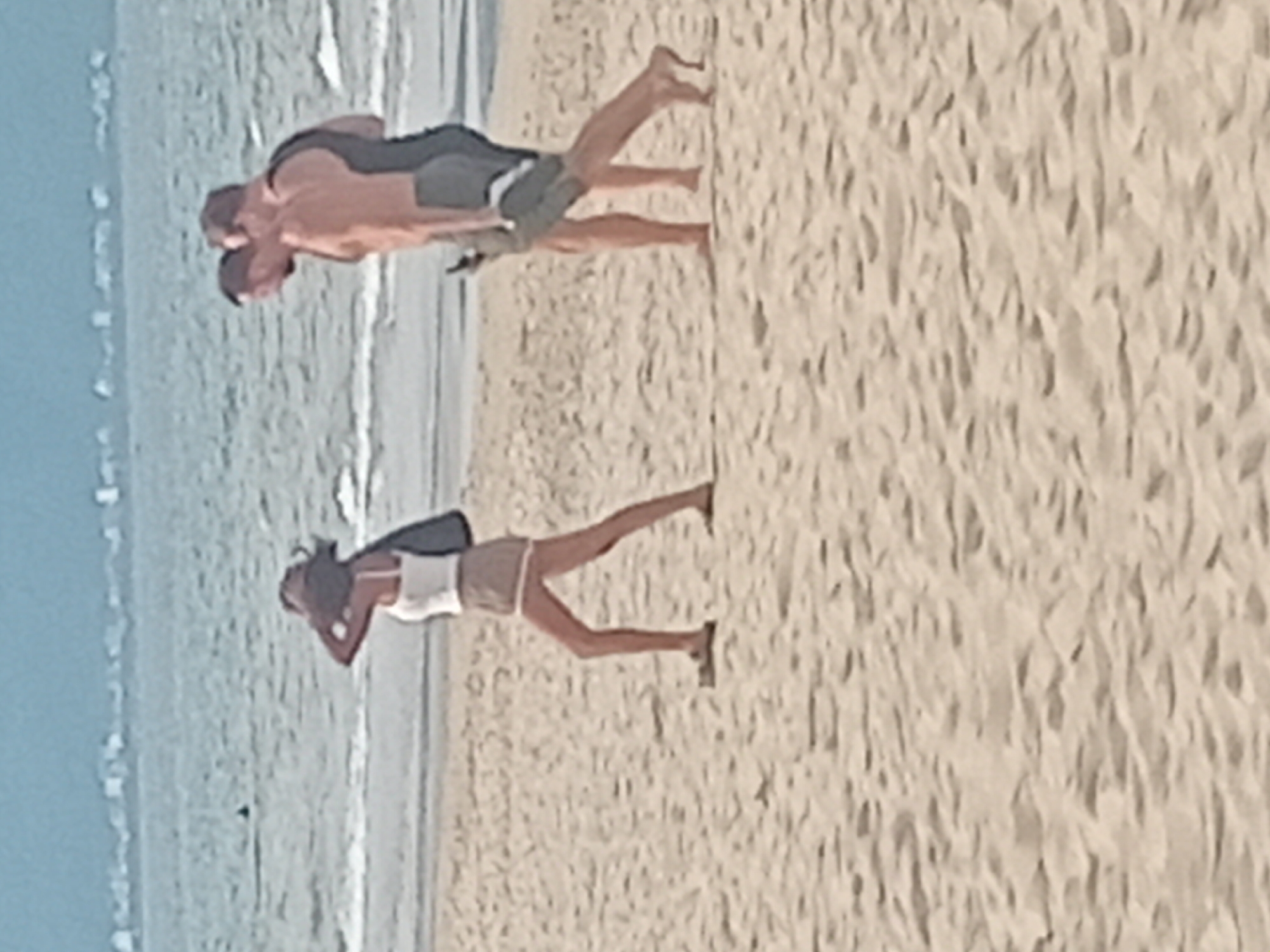 A woman wearing a white tank top, tan shorts, and a wide-brimmed hat walks alone on a sandy beach in Đà Nẵng, Vietnam. She carries a dark bag over her shoulder. In the background, two men walk together further down the beach, one wearing a black tank top and the other shirtless. The ocean is visible behind them with a hazy shoreline in the distance. The image appears to be taken during the daytime under clear skies.