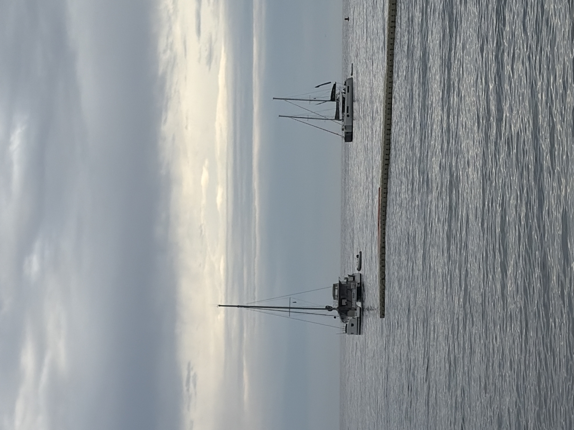 Two sailboats are docked in the water. A barrier of vertical posts is present in the water near the boats. The sky is overcast. The location is likely near Budva, Montenegro, due to the presence of boats and a coastline.