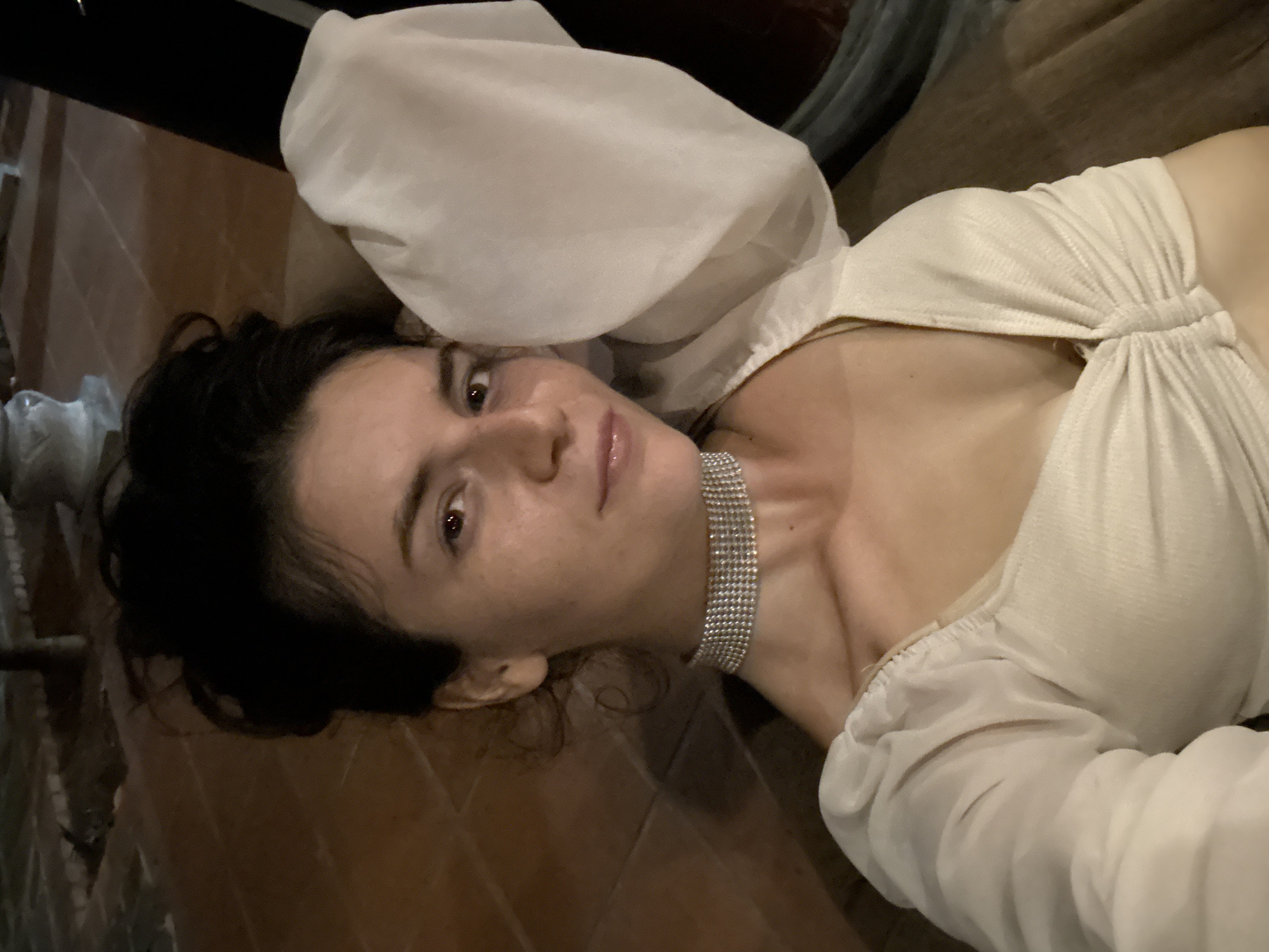 A young woman is lying on a wooden floor. She has dark, wavy hair and fair skin. She is wearing a light-colored, sheer top with large, puffy sleeves, and a multi-row crystal choker. The background includes dark wood flooring and other indistinct items. This scene is presumably in Mũi Né, Vietnam.