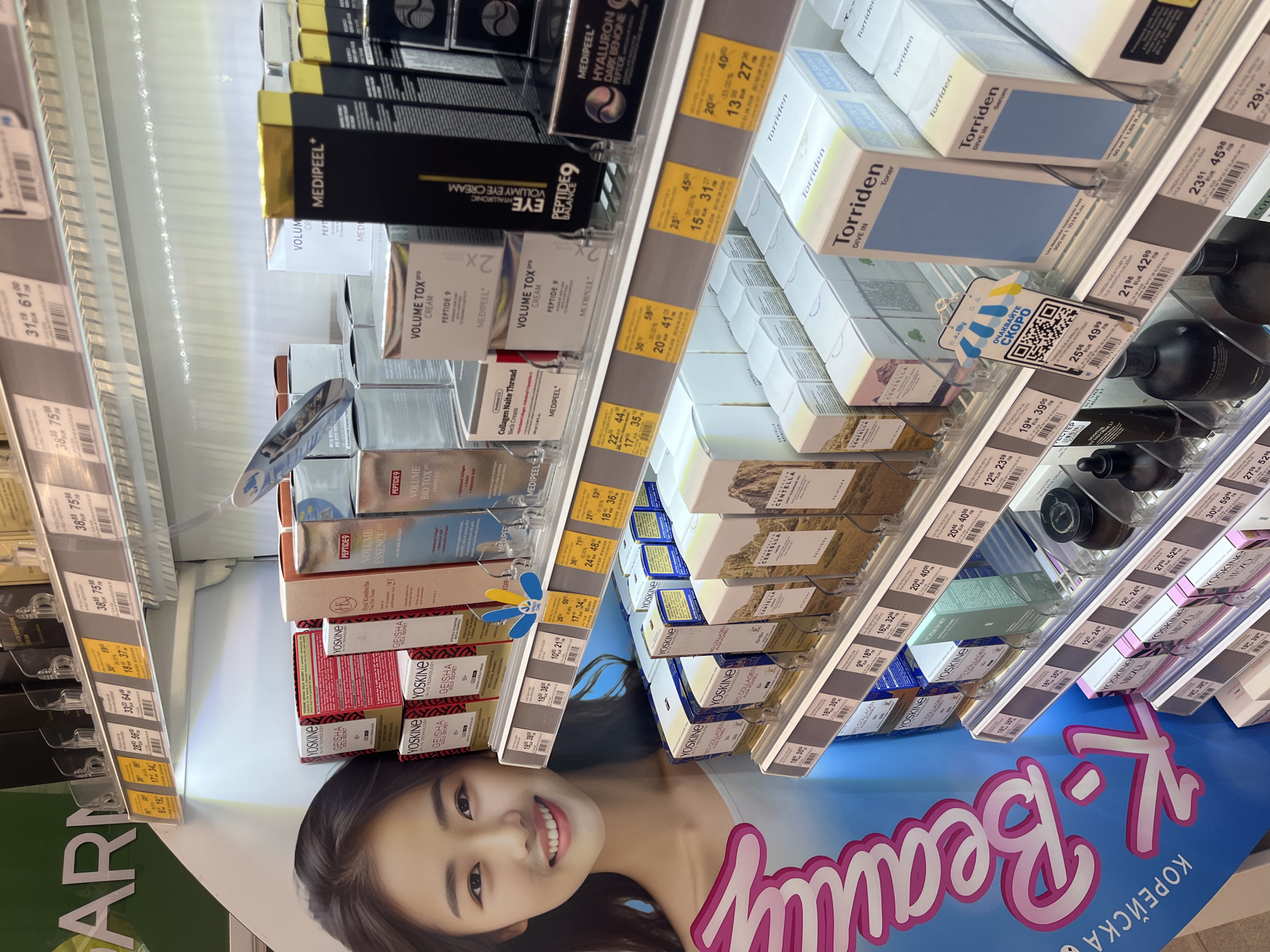 The image captures a brightly lit aisle in a retail store, likely a beauty or pharmacy section, specializing in K-Beauty products. Numerous skincare products are displayed on multiple clear shelves, predominantly in boxed packaging.
The main subjects are the various product lines, including brands like MEDIPEEL, Torriden, YOSKINE, SKINIODA, CENTELLA, and ELIEP. Specific products visible include MEDIPEEL VOLUMY EYE CREAM, VOLUME TOX PRO CREAM, Collagen Naite Thread Neck Cream, Torriden DIVE IN Toner, and Torriden DIVE IN Serum, among others. Many products highlight ingredients such as peptides, hyaluronic acid, centella asiatica, and bio collagen.
At the bottom of the image, a large display features a smiling East Asian woman, reinforcing the "K-Beauty" theme. The text "K-Beauty" and "КОРЕЙСКА" (Koreyska, meaning "Korean") are prominently displayed on this banner.
Price tags are visible for almost all products, showing original prices, discounted prices, and percentage discounts (e.g., 30%, 31%, 33%). Prices are listed in both EUR and лв (Bulgarian Leva), indicating the location is in Bulgaria. One tag also features a QR code with the text "ОЧАКВАЙТЕ СКОРО" (Ochakvayte Skoro), meaning "Coming Soon." The store lighting is artificial and bright, suggesting an indoor commercial setting.