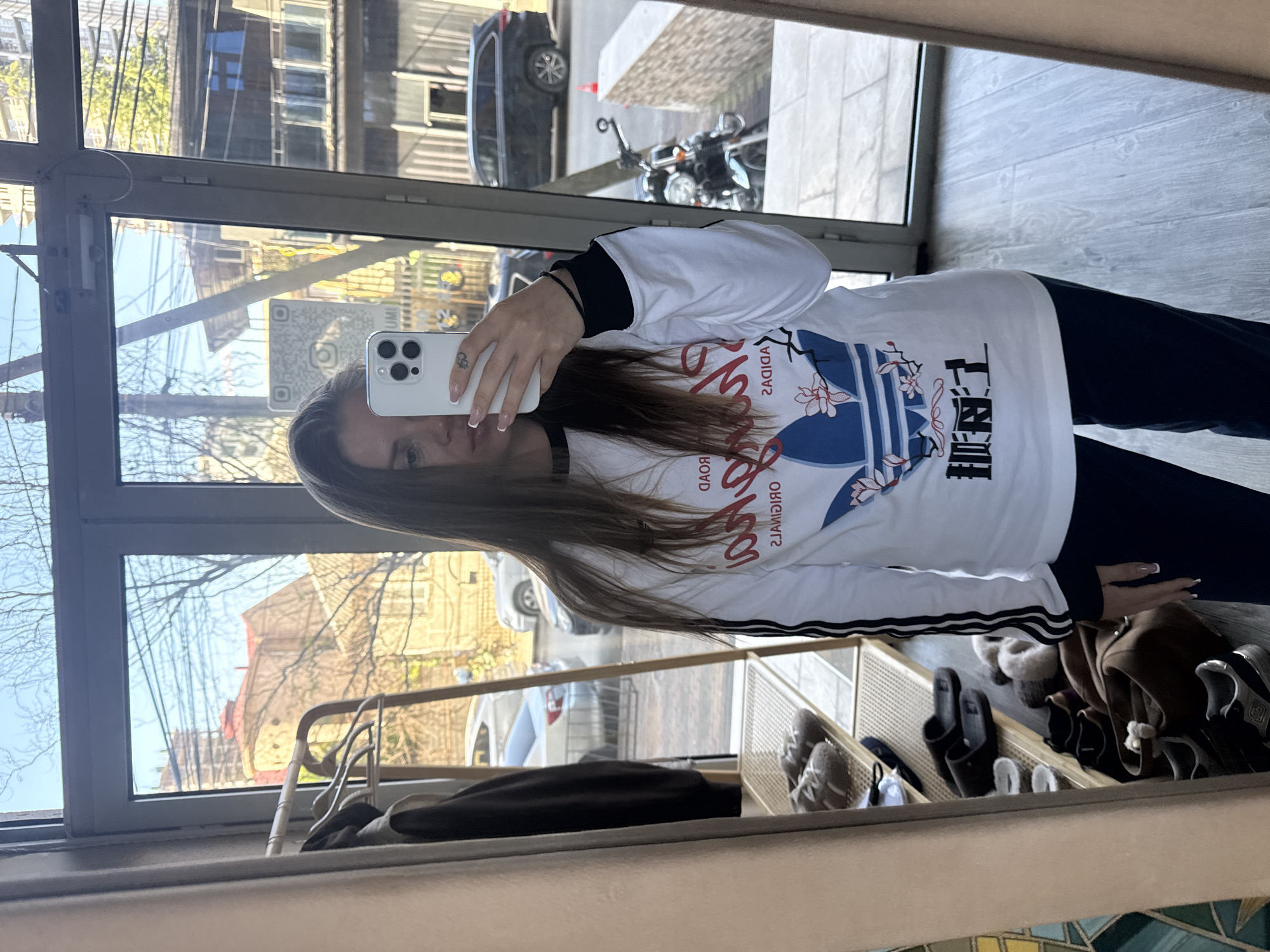 A young woman is depicted taking a mirror selfie indoors, with an outdoor street scene reflected behind her. She holds a white iPhone, likely a 14 Pro model, in her right hand. Her face is partially visible in the reflection, framed by long, straight brown hair. She wears a white long-sleeve shirt featuring a blue Adidas trefoil logo adorned with a floral design resembling cherry blossoms. The shirt also has red text, which appears reversed as "SADIDA" and "SLANIGIRO" in the mirror, indicating "ADIDAS" and "ORIGINALS," along with stylized Japanese characters. Her sleeves have black and white striped accents, and she is wearing dark-colored pants.
The indoor setting appears to be an entryway or hallway. To her right, a shoe rack holds several pairs of shoes, including sneakers and black slippers. A small section of floor at the bottom right shows a mosaic-like pattern with blue, green, and yellow tiles.
The reflection in the large window or glass door reveals a bright, sunny daytime environment. Outside, multi-story urban buildings are visible, alongside trees with mostly bare branches, suggesting a colder season like late autumn or early spring. A dark SUV, a motorcycle, and a small orange cone are parked on the street below. A partially visible QR code sticker with an Instagram logo is affixed to the window or mirror. The bright sunlight and clear sky are notable details. The urban landscape and architectural styles are consistent with a city setting, such as Batumi, Georgia.