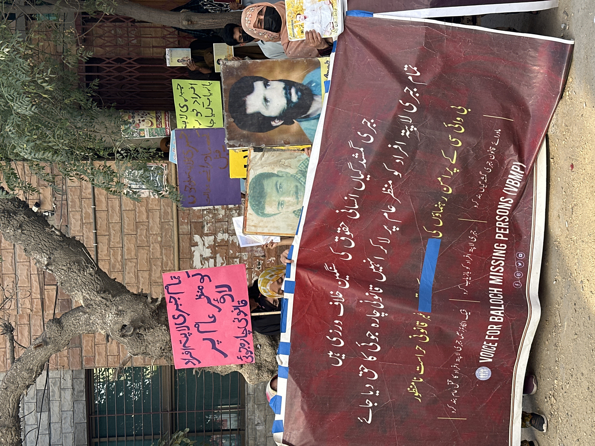 The image depicts a protest or demonstration in Quetta City Tehsil, Pakistan. A large, maroon banner is prominently displayed, bearing text in Urdu and English. The Urdu text translates to "All forcibly disappeared persons should be brought to the public's attention. Illegal detention is unacceptable." The English text on the banner reads "VOICE FOR BALOCH MISSING PERSONS (VBMP)".
Several individuals are visible behind the banner, some holding up photographs of men. One photograph features a man with a beard and dark hair, another shows a younger man. A woman wearing a light-colored headscarf and a black face mask is holding a photograph. Other protesters are partially visible, including a person in a yellow and white headscarf.
In the background, a tree with a rough, gnarled trunk stands in front of a brick building with barred windows. Several signs and posters are attached to the building or on display, including a pink sign with Urdu text and a green sign with Urdu text. The ground is paved with dirt and gravel. The overall scene suggests a gathering focused on raising awareness about missing persons.