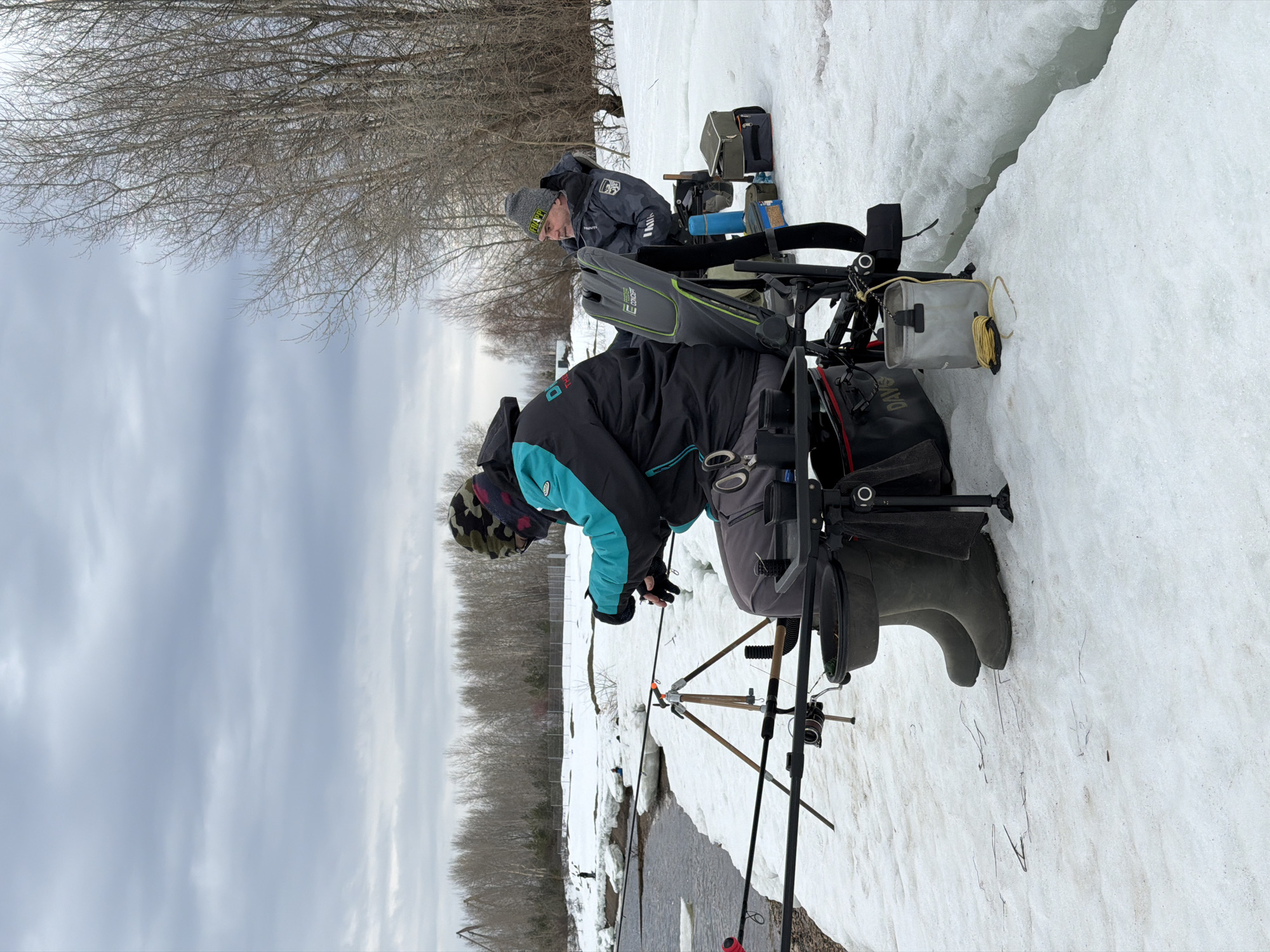 Two men are ice fishing in Noviy, Russia. One man is focused on setting up his fishing rod and equipment. The other is sitting with his own gear. Both are dressed in winter clothing. Snow covers the ground. A river flows in the foreground. Trees are visible in the background. The sky is overcast.
