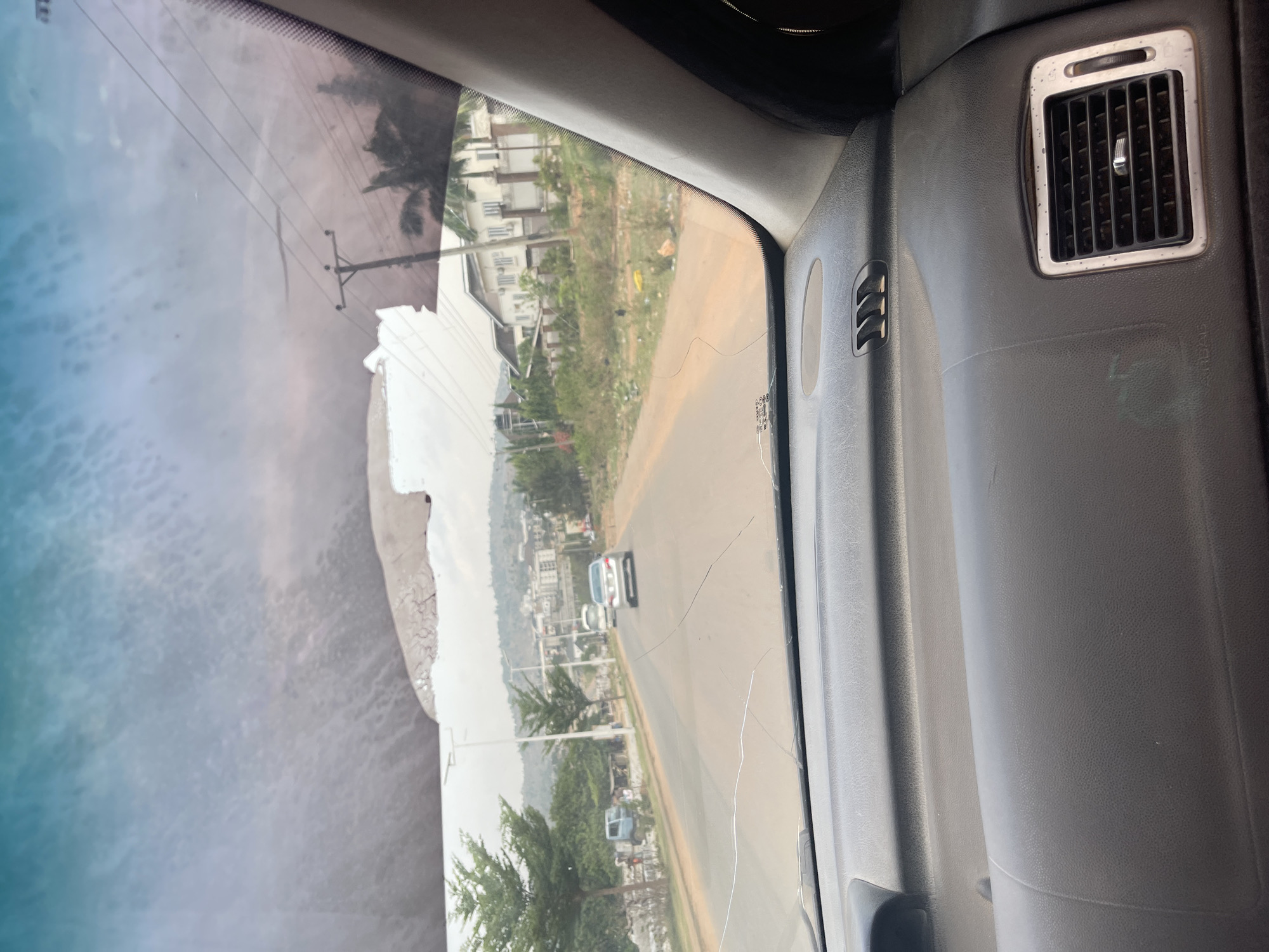 The image presents a view from inside a vehicle, looking out through the front passenger side windshield. The car's gray dashboard and interior plastic trim are visible on the right, featuring a rectangular air vent with silver-colored trim. A faint "AIRBAG" label is discernible on the dashboard.
Through the cracked windshield, an outdoor scene unfolds under what appears to be a clear, bright sky. A paved road stretches into the distance, with a white sedan-like car driving away or parked further down the road. Alongside the road, there's a mix of dry grass, dirt, and some scattered refuse. Utility poles with power lines run parallel to the road. In the background, numerous residential buildings, mostly white or light-colored, are nestled among abundant greenery and trees, suggesting a suburban or semi-urban area. Distant hills or mountains can be seen on the horizon. The top-left portion of the windshield shows reflections of the sky, giving a textured, light blue appearance, and some faint reflections of trees or other structures are also present. The prominent cracks on the windshield are a notable detail. The overall scene suggests a daytime setting in a residential or developing area in Abuja, Nigeria.