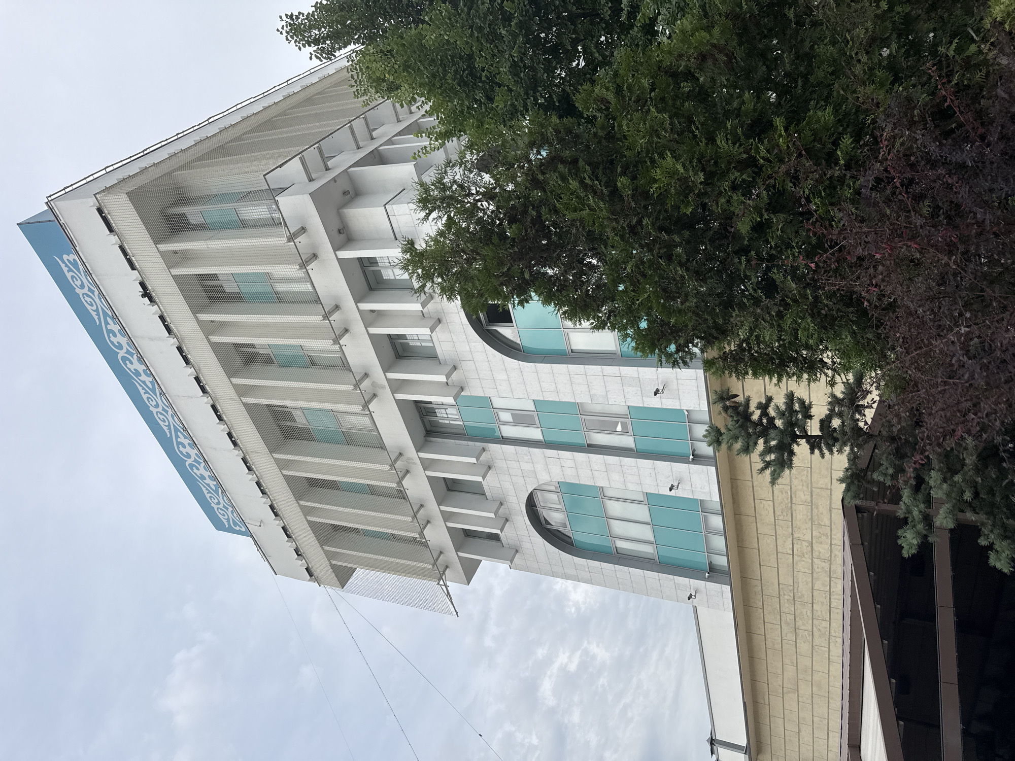 The image shows the exterior of the National Bank of Kazakhstan, located in Almaty, Kazakhstan. The building is a multi-story structure with a modern architectural style, featuring white facades, numerous windows, and a distinctive blue and white patterned sign at the top. Green trees, some with dark foliage, partially obscure the lower sections of the building, indicating an outdoor, possibly urban or park-like setting. The sky is overcast with clouds.